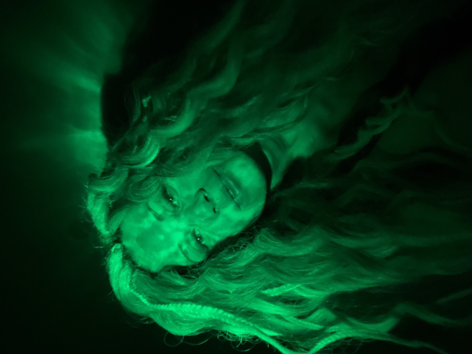 The image depicts a person with long, wavy hair illuminated by a green light source in a dark environment. The person's face is visible, with details like eyes, nose, and mouth discernible despite the green lighting. No specific details linking the image to Rusanovo, Russia are apparent.