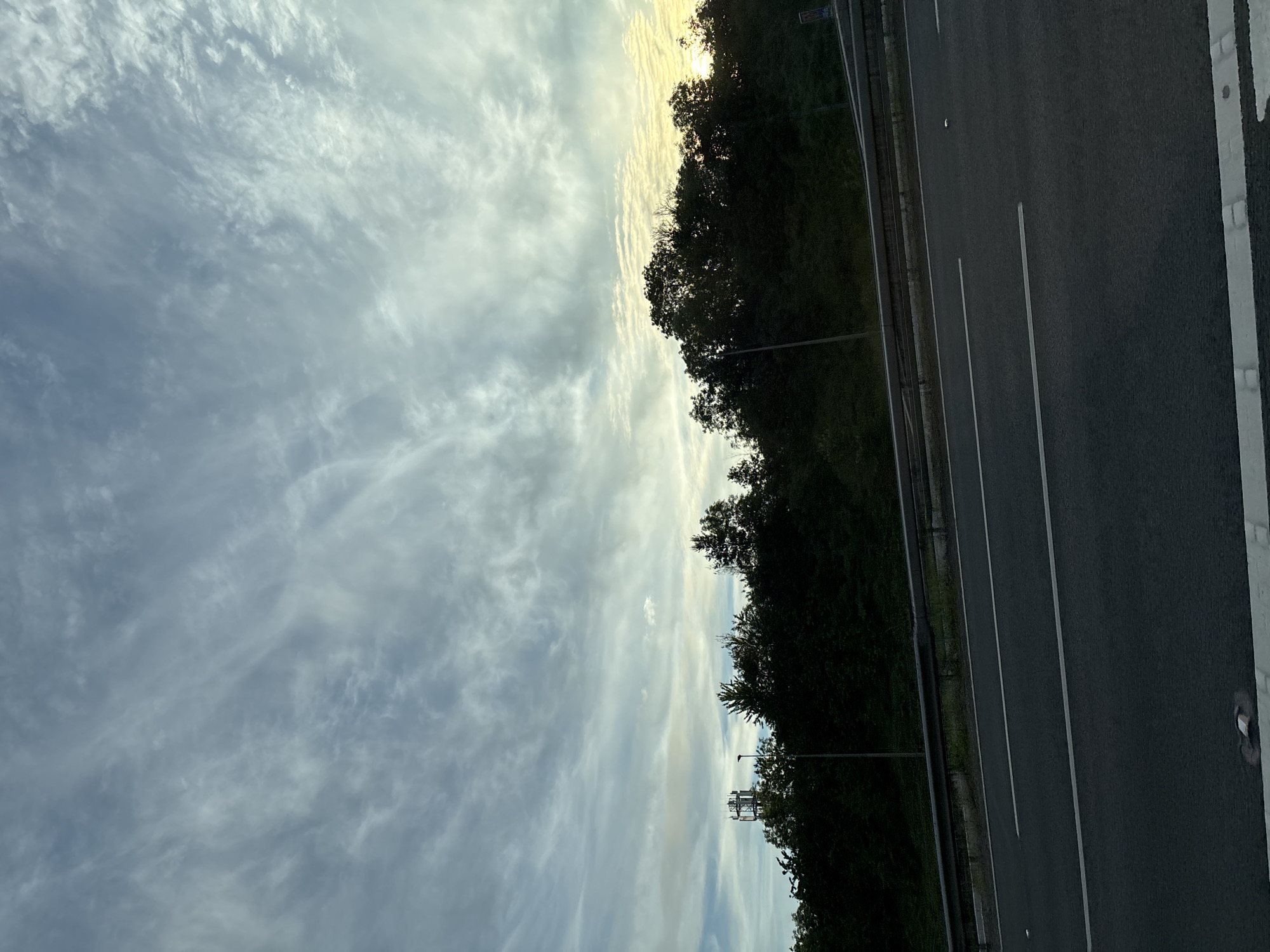 The image shows a rural landscape, likely in South Staffordshire, United Kingdom. A highway runs through the foreground, with lanes marked by white lines. Beyond the highway is a dense line of dark green trees, with a single street light visible amidst them. In the distance, to the left, a tall, metal telecommunications tower stands above the treeline. The sky above is filled with wispy, white clouds against a pale blue background. The lighting suggests it is either early morning or late afternoon, with a hint of golden light on the right side of the horizon. There are no people or vehicles visible in the image. The overall impression is one of natural beauty combined with infrastructure.