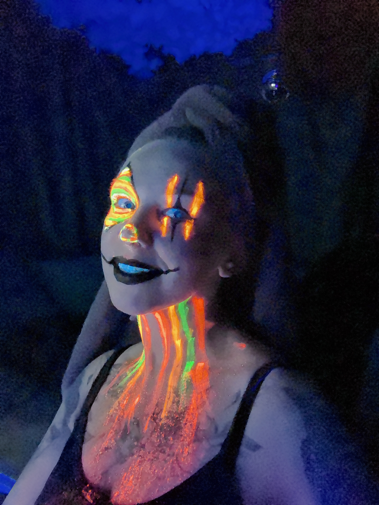 A person with clown makeup stands in a dark environment, presumably indoors. The makeup is brightly colored with orange, yellow, and green patterns around the eyes and on the neck. The mouth is painted black. The person is wearing a dark-colored tank top. The background is dark, and the location is not identifiable as Noviy, Russia, or any other specific city.