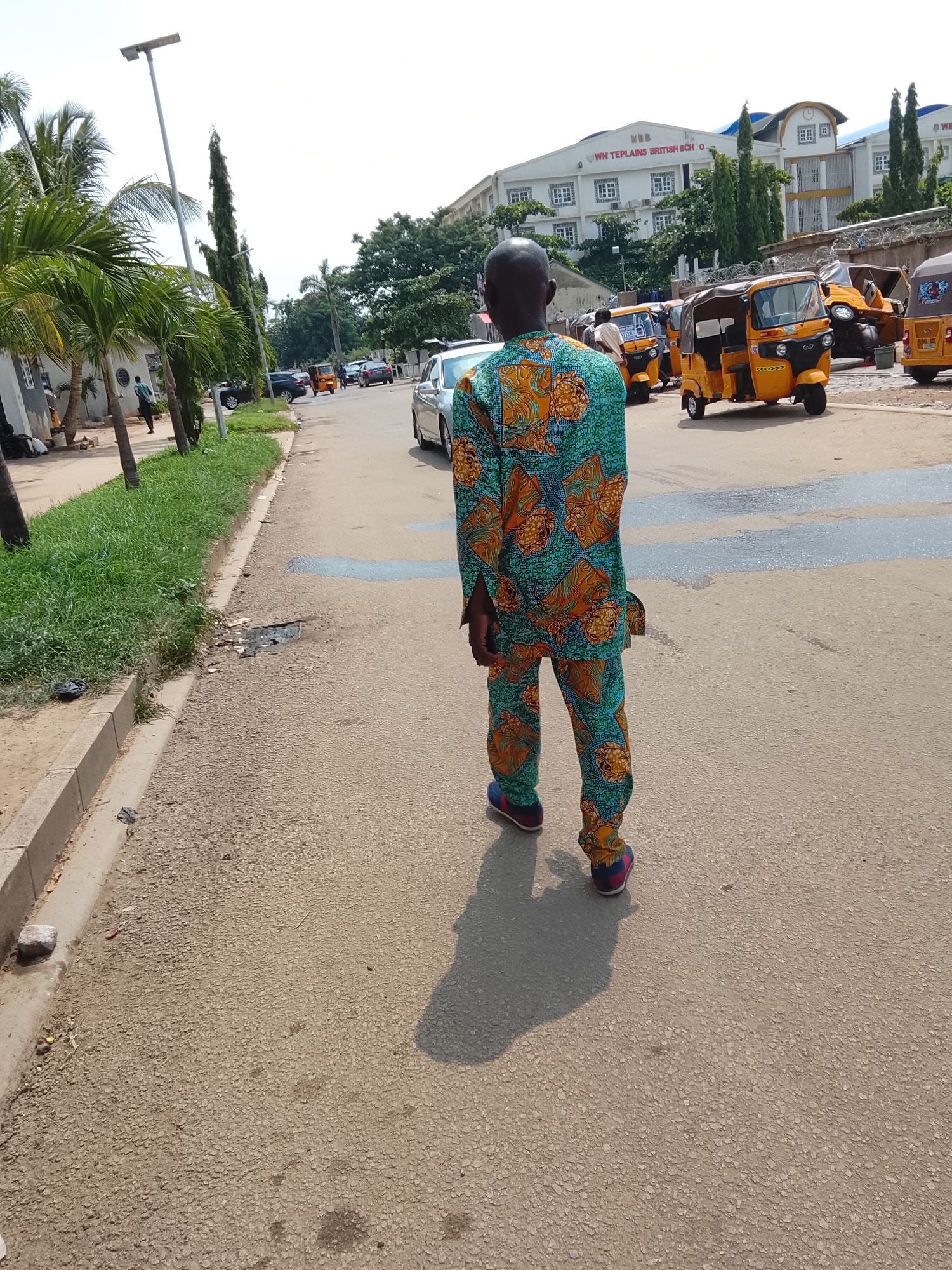 In Abuja, Nigeria, a man wearing a vibrantly patterned green and orange dashiki walks down a paved street. The man is facing away from the camera, his shadow stretching out before him. On the left side of the street, a grassy verge lines the sidewalk, with palm trees and other vegetation present. In the background, several yellow auto rickshaws are parked. Behind the auto rickshaws, a large, multi-story building with white and yellow facade and numerous windows is visible, bearing the sign "WN TEPLAINS BRITISH SCH O." The sky is clear and bright, suggesting a sunny day. Several cars are also visible in the distance, along with other pedestrians.