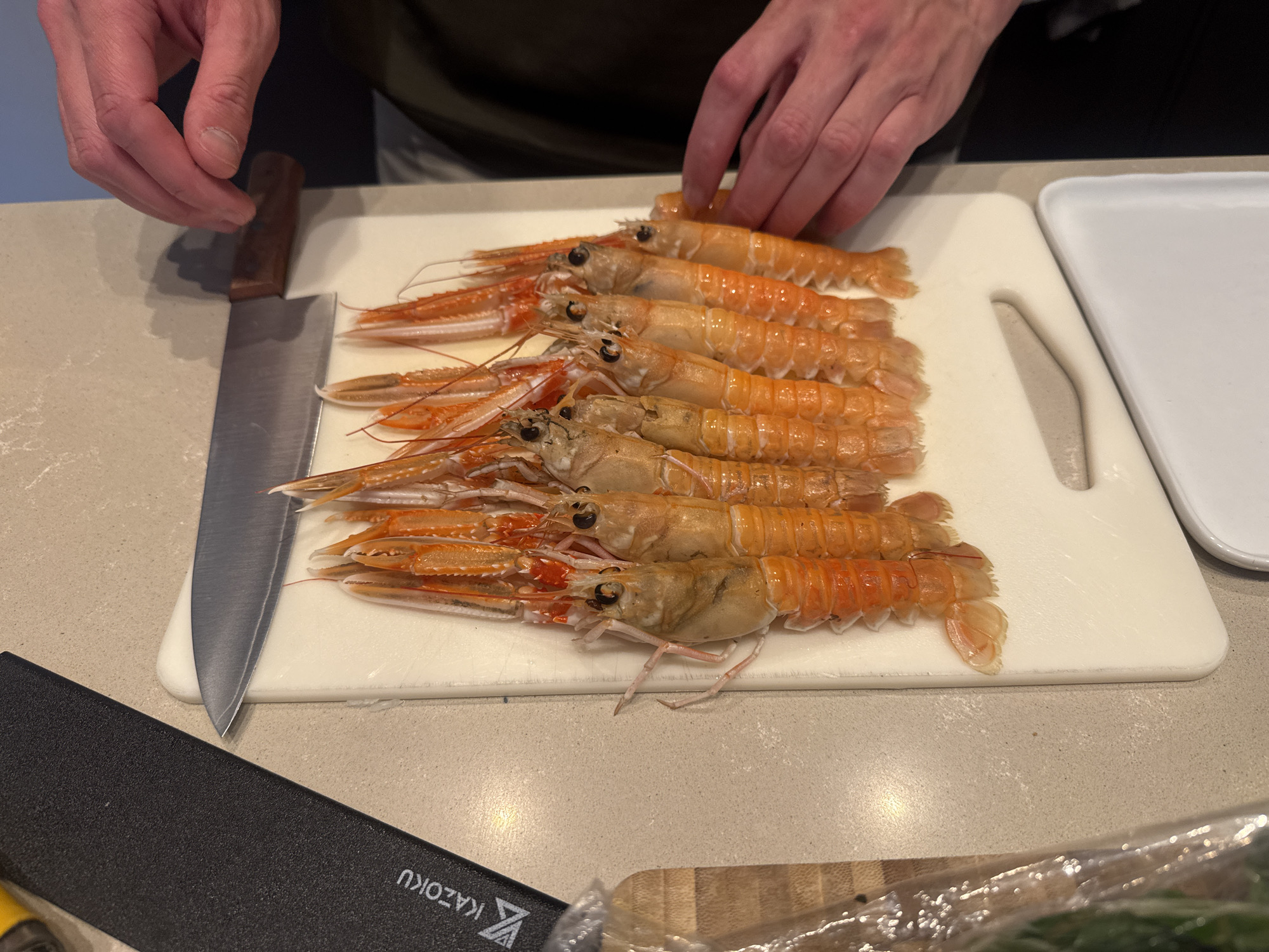 A close-up shot depicts a person preparing langoustines on a white cutting board. The individual is seen touching the row of crustaceans, which are arranged horizontally on the board. A kitchen knife rests to the left of the cutting board, alongside a sheath or protective cover for the knife. Another white, rectangular plate is located to the upper right of the cutting board. The scene appears to be set on a countertop, and it includes a knife bearing the brand name "KAZOKU". The specific location is not discernible, and there is no indication that this photo was taken in Amsterdam, Netherlands.
