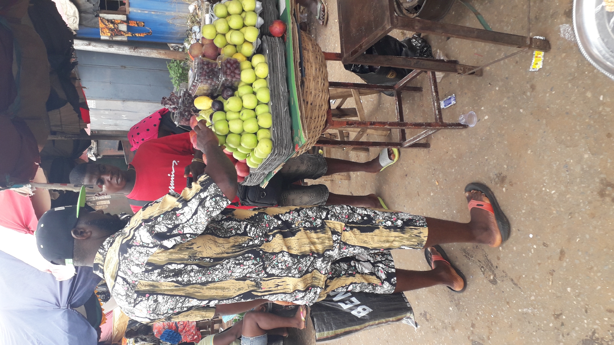 The image shows a street scene in Nkpor, Nigeria. Two men are visible, one wearing a black cap and a patterned shirt, the other in a red shirt with a black dotted design. The man in the cap is standing near a fruit stand. The fruit stand is stocked with green apples, grapes, and some red fruit. The fruit stand is on wheels. There is a metal structure with a wooden base. The ground appears to be concrete.