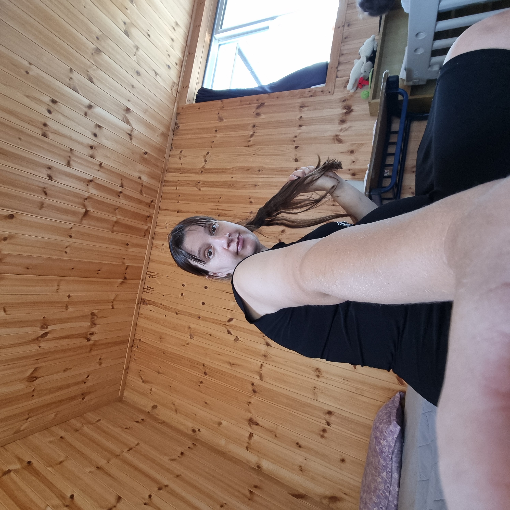 The image depicts a woman in a black top, seated on a bed. She is holding a strand of her brown hair in her right hand and looking towards the camera. The background consists of wooden paneled walls and ceiling, indicative of a room with a rustic or cabin-like interior. A window with blue curtains is visible on the right side of the frame, letting in natural light. On a shelf to the right of the window, several objects are present, including a white stuffed animal and a small green ball. Part of a child's crib or playpen is also visible in the lower right corner of the image. The woman is positioned in the center of the frame, with her left arm and shoulder prominently in the foreground, suggesting a selfie-style photograph. The setting is indoors.