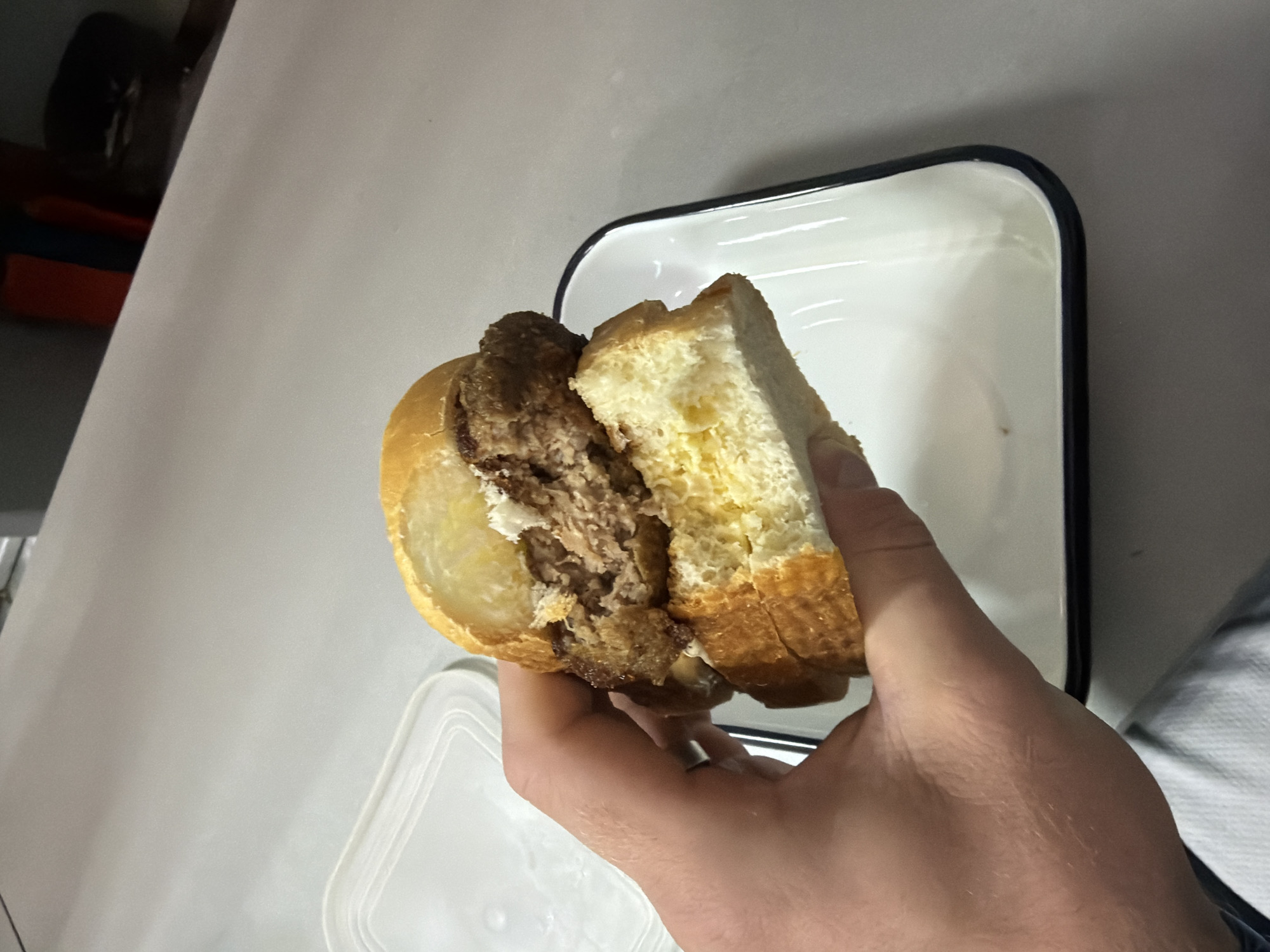 An indoor, slightly angled overhead shot captures a hand holding a large sandwich or burger. The prominent subject is a golden-brown bun, split open to reveal a substantial, dark, and coarsely textured meat patty inside, likely ground beef. The interior of the bun appears lighter, with a yellowish hue. The hand holding the sandwich belongs to an adult, with visible skin texture and some light hair, indicating an active grip on the food.
In the upper right portion of the frame, a white rectangular enamel plate with a distinct dark rim rests on a light-colored surface, appearing empty. Partially visible in the lower left is a translucent white plastic container lid, showing subtle ridges. The background in the upper left features blurry, dark shapes, and indistinct colorful objects (orange, blue, black), suggesting storage or furniture within the room. The overall setting appears to be a casual dining or preparation area, possibly a kitchen.
The lighting throughout the image is even, suggesting artificial illumination or diffused natural light. While the location context is Chaikovsky, Russia, there are no specific visual cues within the image to confirm this. No text is visible in the frame, and no particular emotion is conveyed by the subject's hand.