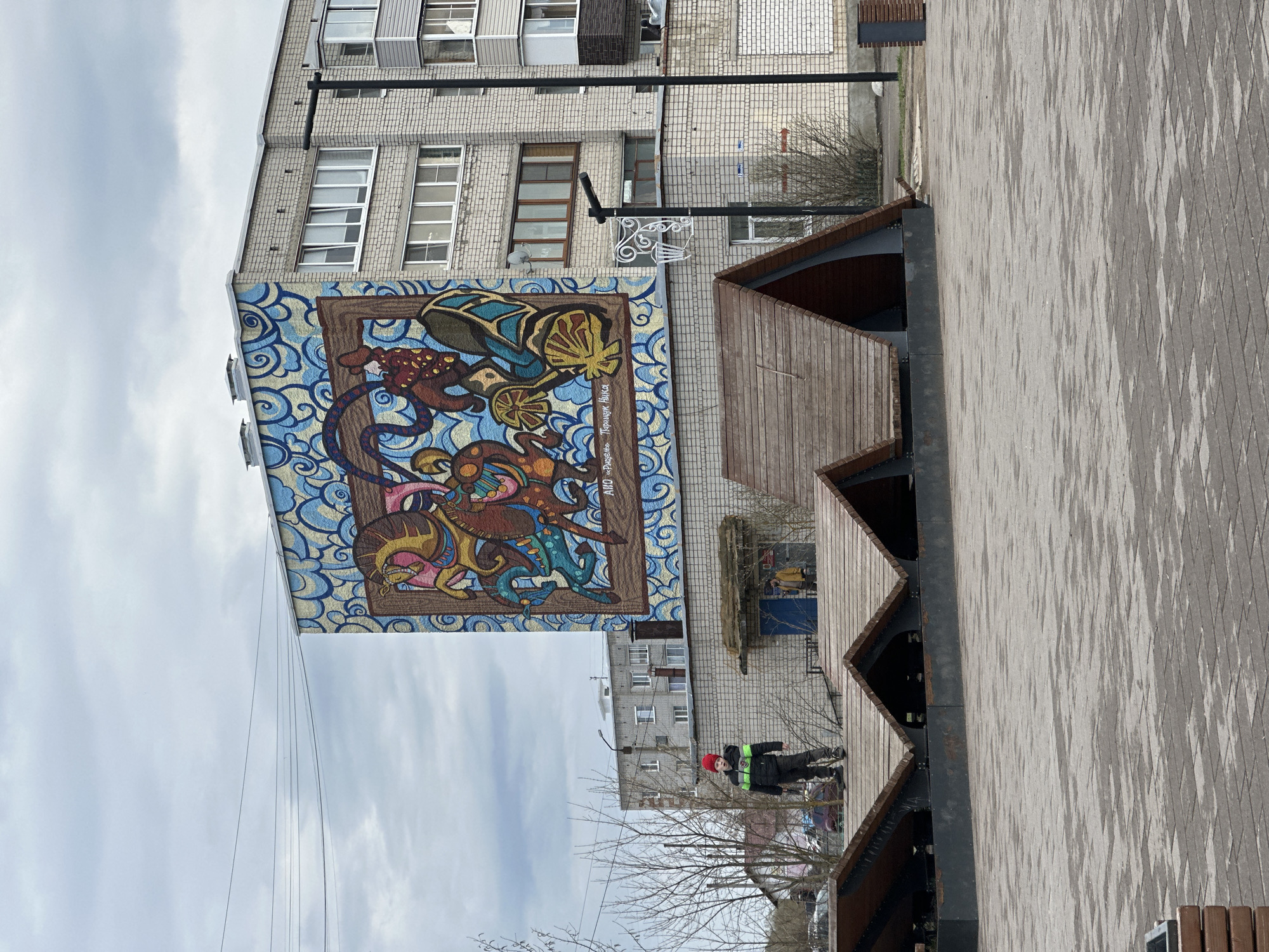 The image depicts a building in the city of Pokrov, Russia, featuring a large mural on its side. The mural, framed by a brown border, showcases an abstract and colorful artwork against a blue and white swirling background. The building itself is constructed of light-colored bricks and has multiple windows. In the foreground, there's a walkway with wooden structures resembling roof sections. A person wearing a red hat and dark clothing with reflective stripes is visible walking on the walkway. The ground is paved with gray bricks in a patterned design. The sky is overcast with light clouds. Power lines are visible stretching across the top part of the image.