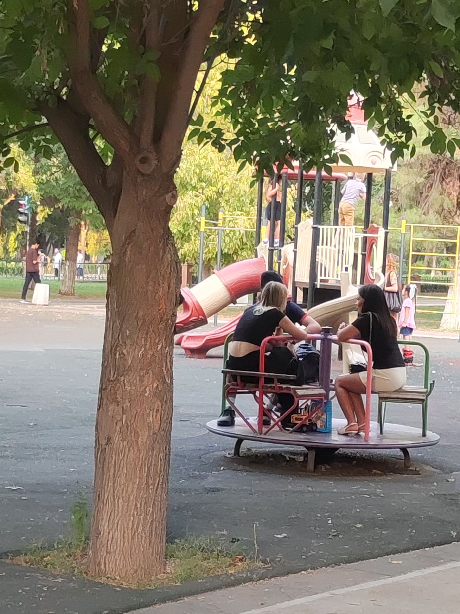In Tashkent, Uzbekistan, a merry-go-round at a playground has three individuals seated on it, facing inwards. One person on the left, partially obscured, wears a black top and dark pants. In the center, another person in a black top and dark pants is visible. On the right, a person in a black top and a light-colored skirt is seated, with their legs extended and wearing white sandals. A red and white plastic slide is visible to the left of the merry-go-round, connected to a playground structure. In the background, other children are playing on the playground equipment, and a tree with green foliage stands prominently in the foreground on the left. The ground surrounding the playground appears to be asphalt or a similar paved surface.