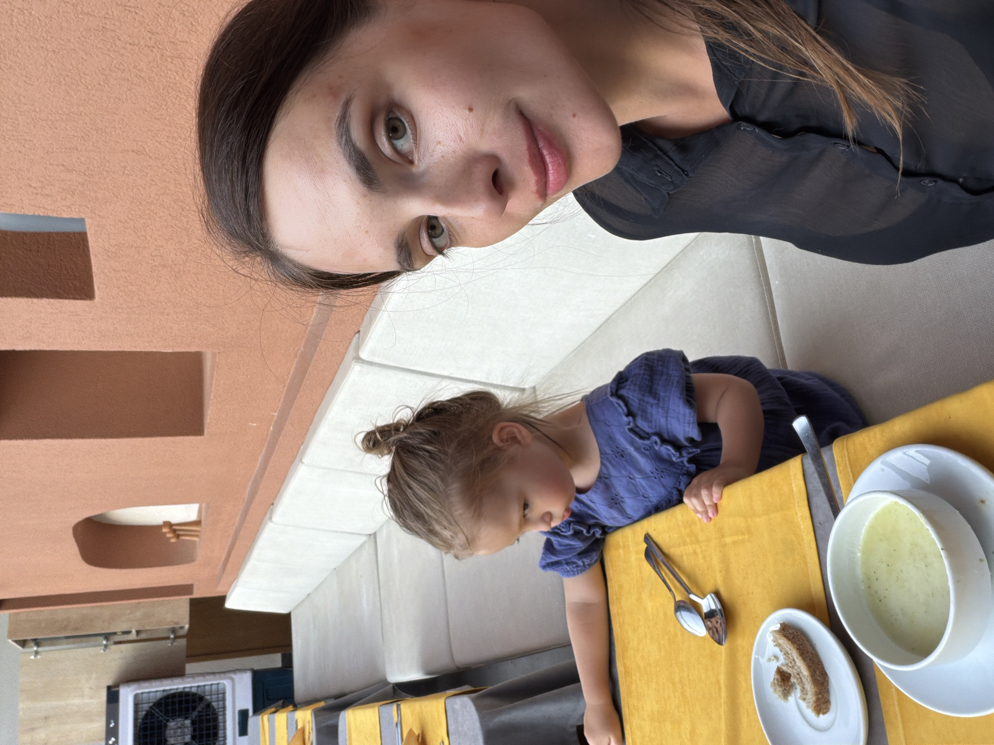 A woman and a young girl are seated at a table on a patio. The woman is in the foreground, taking a selfie. She has dark brown hair and is wearing a sheer black blouse. The girl is seated to the woman's left, wearing a blue dress. She is looking down at a bowl of soup on the table, which also has a piece of bread on a plate and two spoons. The table is covered with a yellow tablecloth. In the background, there are outdoor benches with a light-colored upholstery, and a wall with an orange stucco finish. To the left, a white industrial fan is visible. The scene appears to be outdoors, possibly at a restaurant in Fethiye, Turkey, given the architectural style and the presence of outdoor seating.