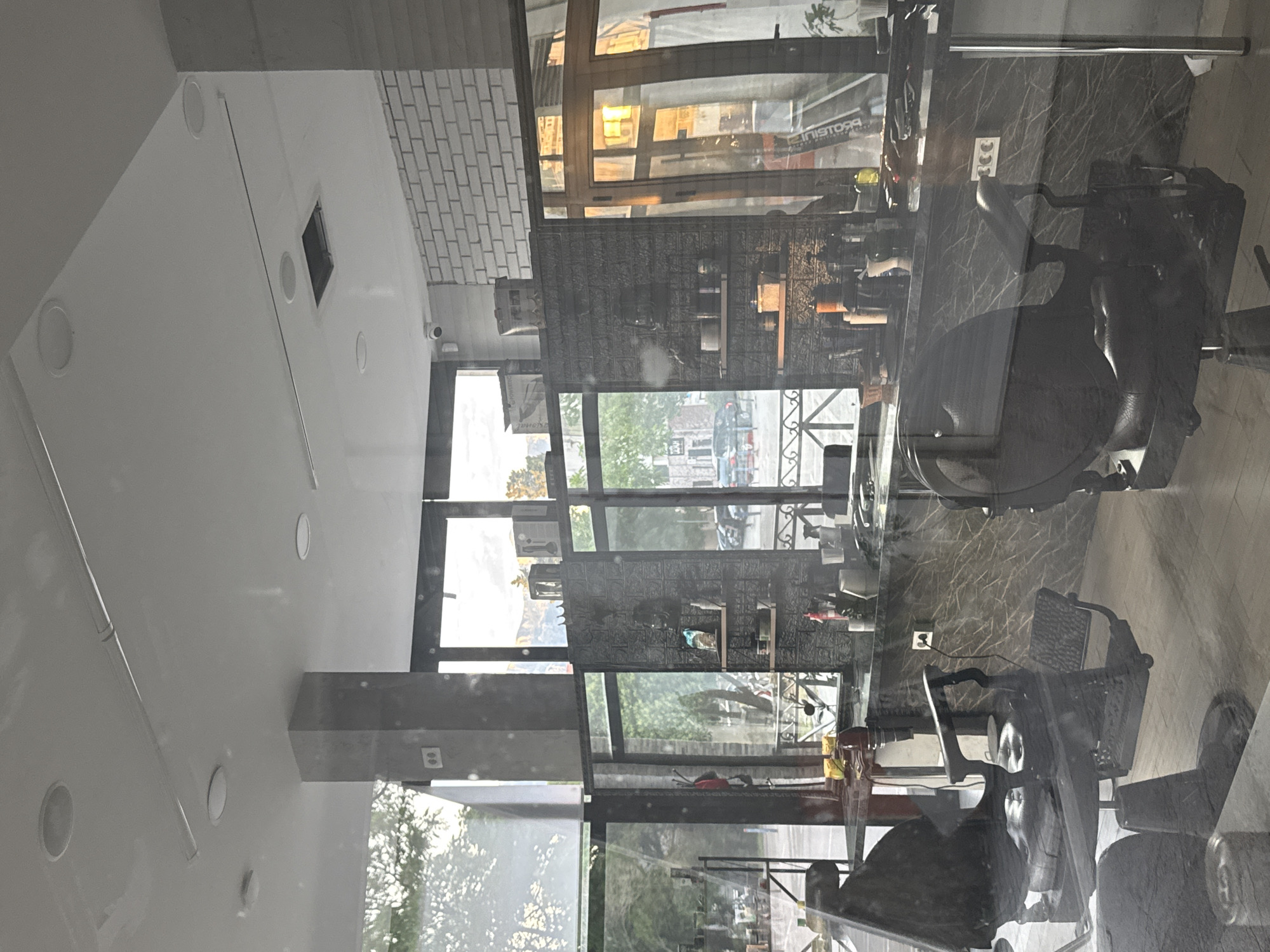 The image depicts the interior of what appears to be a barbershop, reflected in a large window that overlooks a street. The space features white walls with embedded lighting fixtures, and a section of brick wall. The reflection in the window shows the street outside, with buildings and trees visible, suggesting a daytime scene. Inside, barber chairs and countertops are visible, providing context to the shop's function. Based on the image, it is difficult to determine the precise location in Bar, Montenegro, but the scene shows a typical urban setting with commercial activity.