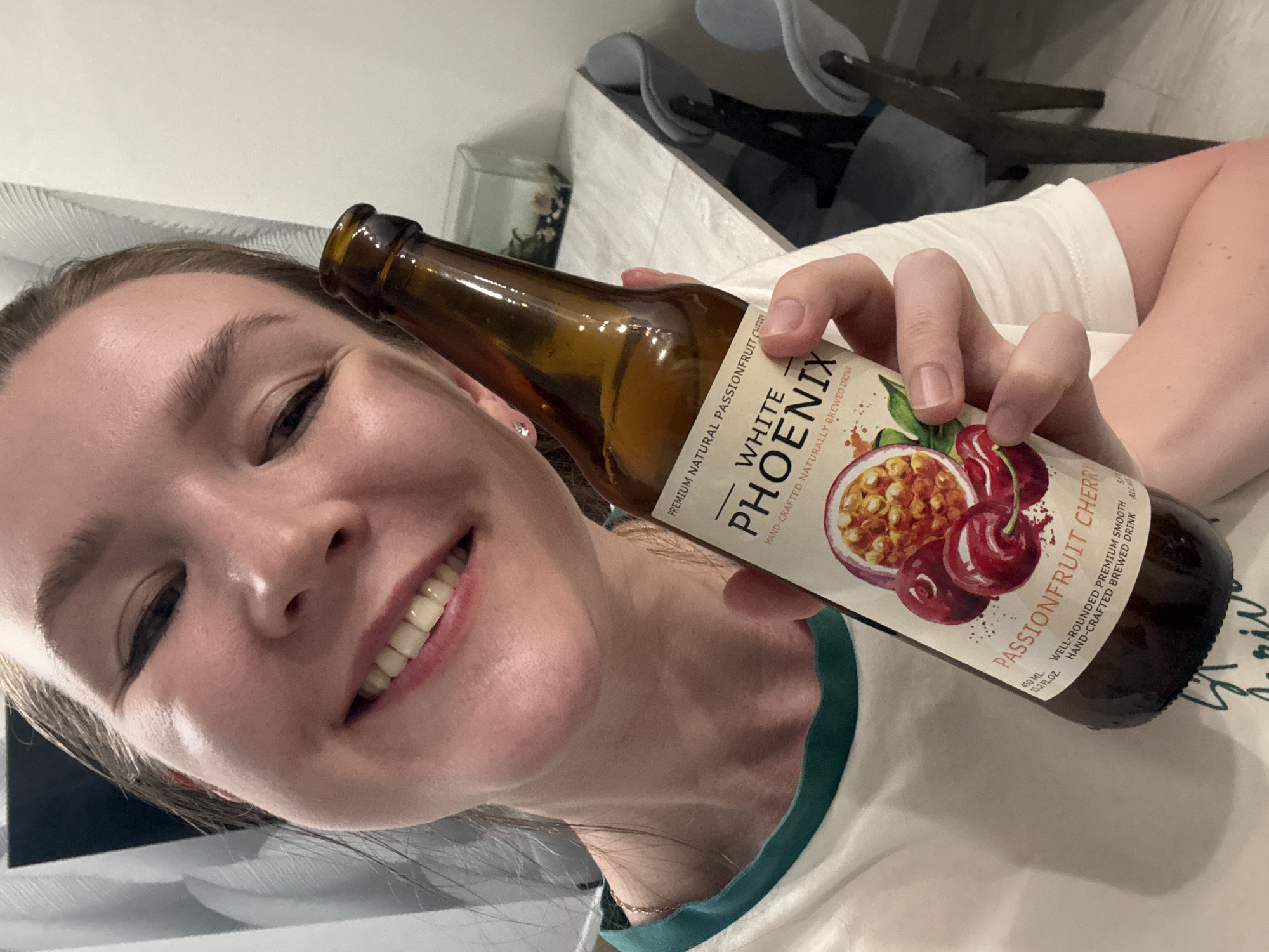 A woman is holding a bottle of "White Phoenix Passionfruit Cherry" drink. The woman is wearing a white T-shirt with a green neckline. In the background are chairs. The city of Izhevsk, Russia, is not explicitly visible in the image and can not be determined.