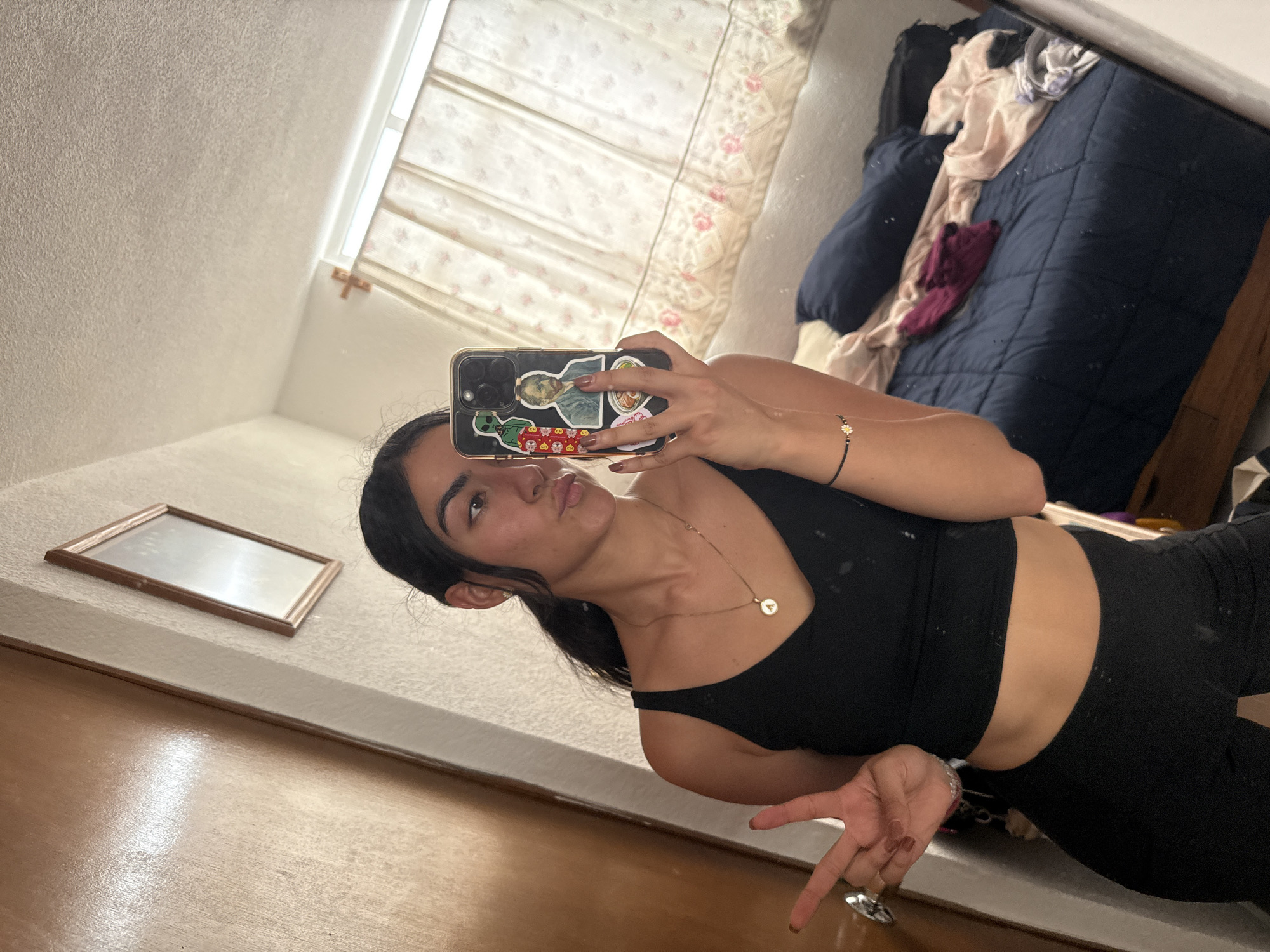 A young woman takes a mirror selfie indoors, likely in a bedroom in Puebla, Mexico. The image is captured from a tilted, diagonal perspective.
The subject, a woman with dark hair, is wearing a black sports bra or crop top and matching black leggings. She poses with a slight pout, looking towards the camera in the mirror. Her right hand holds a black smartphone with a multi-lens camera, which is adorned with several stickers, including a classical bust, cartoonish red skulls, and a bowl of noodles. Her fingernails are painted a muted brown/mauve. On her right wrist, she wears a delicate beaded bracelet with a white daisy charm, and around her neck, a thin gold chain with a small, circular pendant (possibly an initial 'F'). Her left hand, with similarly painted nails, is visible in the foreground, making a peace sign.
The background reveals a personal space with natural light filtering through a window fitted with light-colored, floral-patterned blinds or curtains. A messy bed with a navy blue comforter and various clothes (pink/beige and purple/maroon) is seen behind her. The walls are light-colored and textured, and a wooden floor is visible. A small, empty wooden picture frame rests against the wall in the mid-ground. The overall impression is a casual, lived-in room during daylight hours. No discernible text is clearly visible other than potential symbols or a single initial on the necklace.