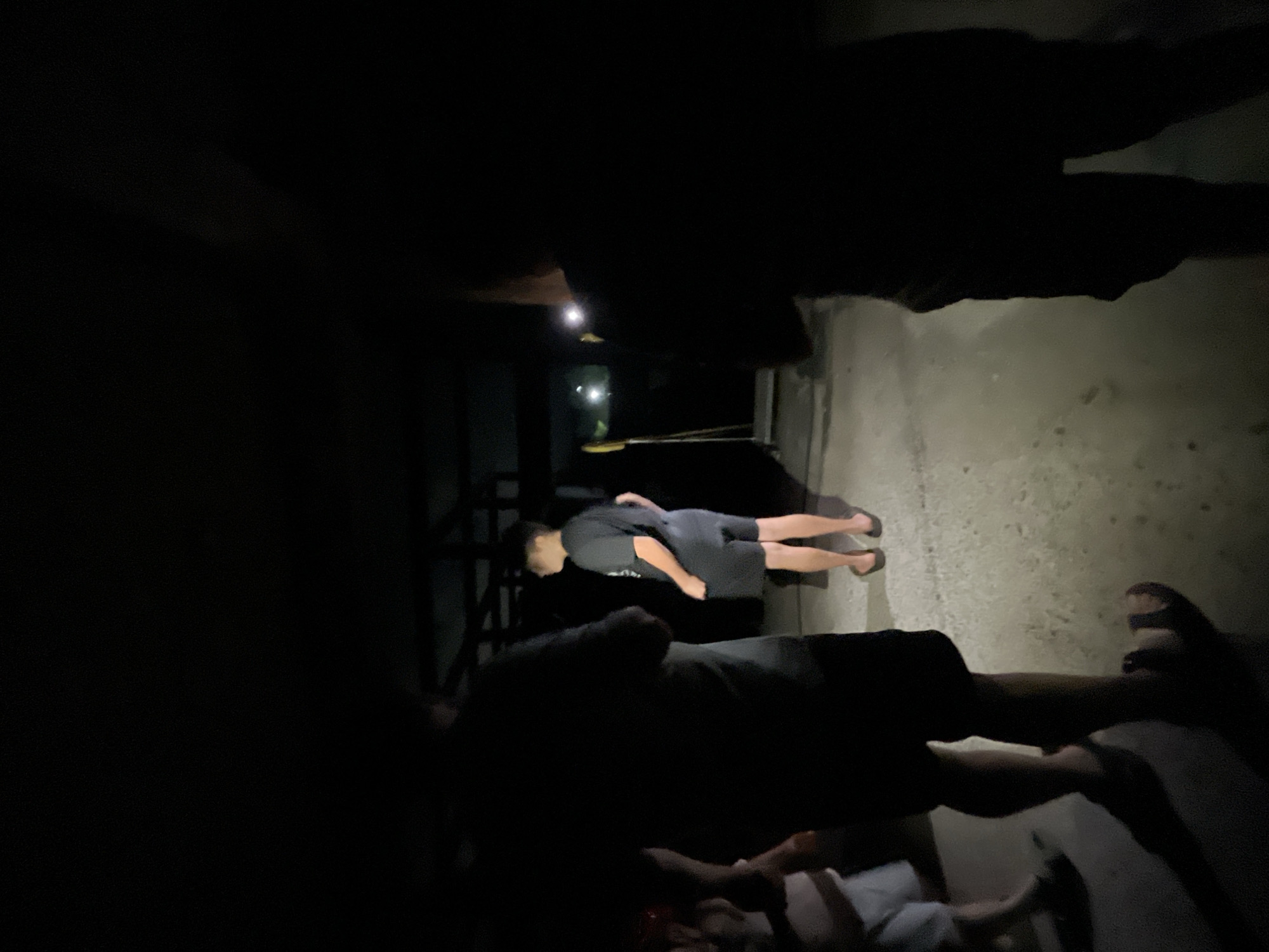 A group of individuals are gathered at night in an outdoor, partially covered area. The lighting is dim, with some artificial lights visible in the background, casting long shadows. In the foreground, the legs of two people are visible, one wearing shorts and sandals, the other obscured. In the midground, a person wearing a dark t-shirt and shorts stands facing away from the camera, with another individual partially visible to their left. The ground appears to be concrete or asphalt. In the background, structures suggest a covered patio or building, with some objects like poles and what looks like a broom leaning against a wall. The overall impression is that of a casual gathering in an urban or semi-urban outdoor setting. No specific landmarks in Poti, Georgia are identifiable.