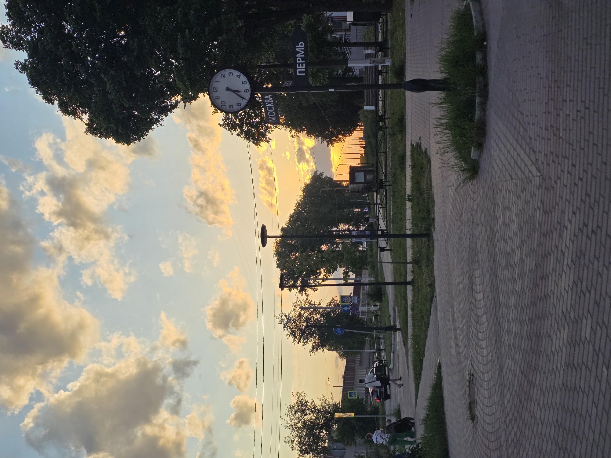 The image shows a street in Chaikovsky, Russia. A sign with the names of cities "Moscow" and "Perm" is visible, along with a clock indicating a time just after 8:00. A sunset is in the background. The foreground features a paved walkway with grass. In the distance, people are standing near a car. Overhead, utility lines stretch across the sky.