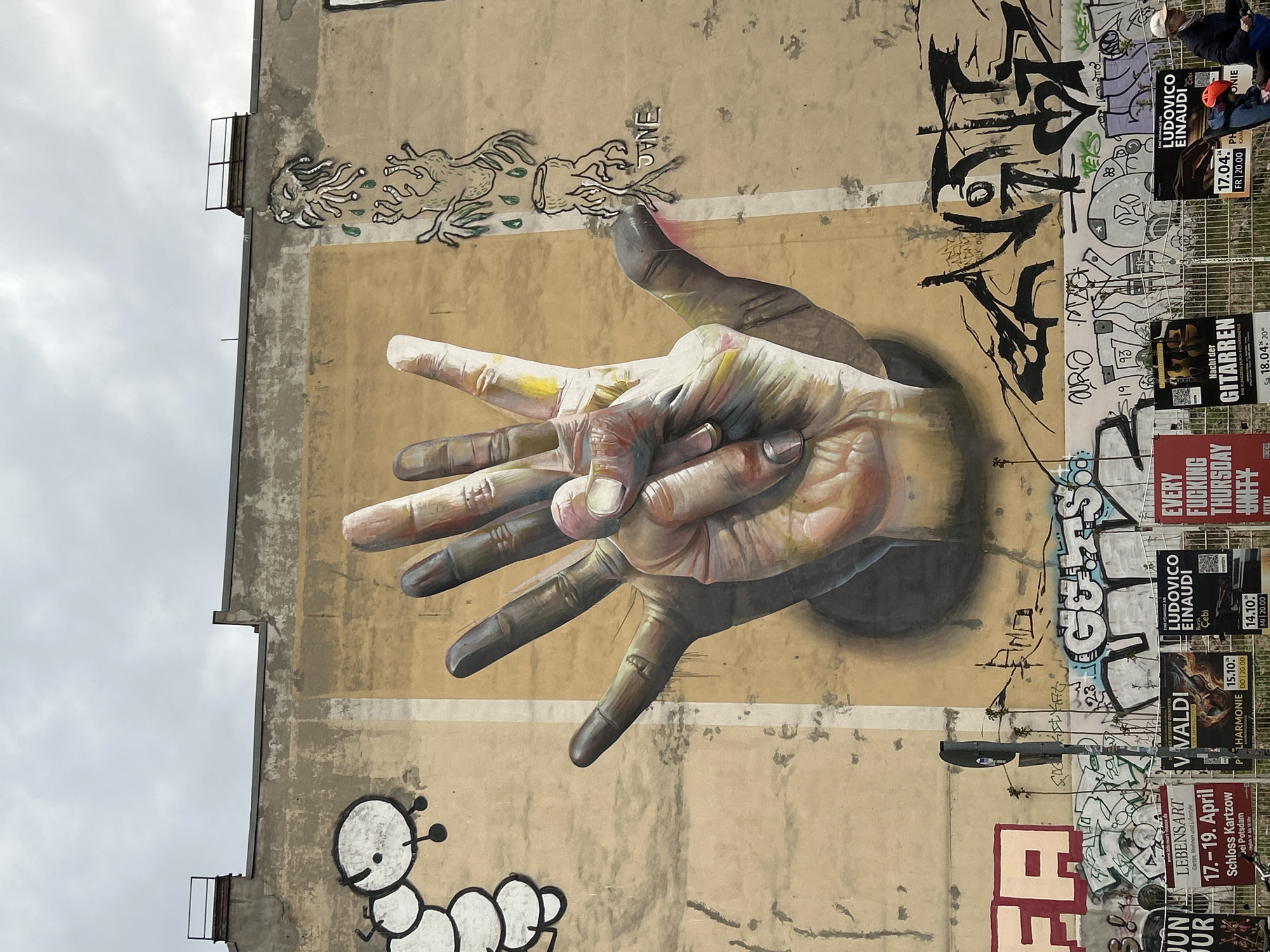 A large-scale mural adorns the side of a beige urban building, capturing a vibrant street art scene. The central feature is a highly realistic, three-dimensional painting of two hands. A lighter-toned hand, showing colorful paint smudges, is held up with splayed fingers in a gesture that resembles a peace sign or counting. This hand appears to emerge from a dark, circular opening in the wall, slightly in front of another, darker-toned hand which also extends outward.
Surrounding the main mural are various styles of graffiti. To the upper left, two stylized, tentacled creatures are depicted, seemingly interacting or spewing liquid. Below the mural, a whimsical black and white caterpillar with large eyes crawls along the wall. The right side and upper portions of the wall are covered in numerous graffiti tags and lettering, including visible words like "JANE," "GUS ORO," "MA," "FA," and the year "1993."
The rightmost section of the wall is heavily adorned with event posters, suggesting a lively cultural calendar. Visible events include concerts by "LUDOVICO EINAUDI" (dated 14.10. and 17.04.), "Nacht der GITARREN" (Night of Guitars) on 18.04., and "V VALDI" (Vivaldi) on 15.10. There's also an advertisement for "LEBENSART Garten, Wohnen und Lifestyle" (Lifestyle: Garden, Living and Lifestyle) from 17.-19. April at Schloss KartzoW in Potsdam, and a poster for "EVERY FUCKING THURSDAY KITKAT," likely referring to the famous Berlin club.
A few partially obscured people can be seen near the posters on the far right, possibly observing the art or passing by. The building features a metal fire escape or ladder on its left side. The sky is overcast and cloudy, indicating daytime. The scene vividly portrays an eclectic urban environment, characteristic of street art prevalent in cities like Berlin.