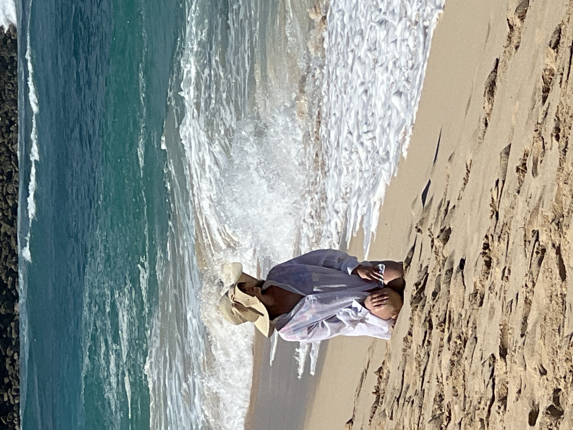 This brightly lit photograph captures a serene beach scene in Kuta Selatan, Indonesia, under clear, sunny skies. In the foreground, positioned on the right, a person is seen relaxing on the light brown sand, seemingly reclining or sitting, facing towards the ocean. They are wearing a wide-brimmed, light-colored hat and a loose, light-colored long-sleeved shirt, possibly white with faint plaid patterns. Their hands are gently resting in their lap or clasped together.
The vibrant blue-green ocean dominates the left side and background of the image, with foamy white waves actively breaking and receding onto the shore, creating a dynamic edge between the water and the sand. The wet sand near the waterline shows intricate patterns from the wave action, while the drier sand further up the beach also displays textures and minor undulations. Faint dark, possibly rocky, formations can be seen along the distant shoreline to the far left. The overall impression is one of peaceful leisure on a sunny tropical beach. There is no visible text in the image.