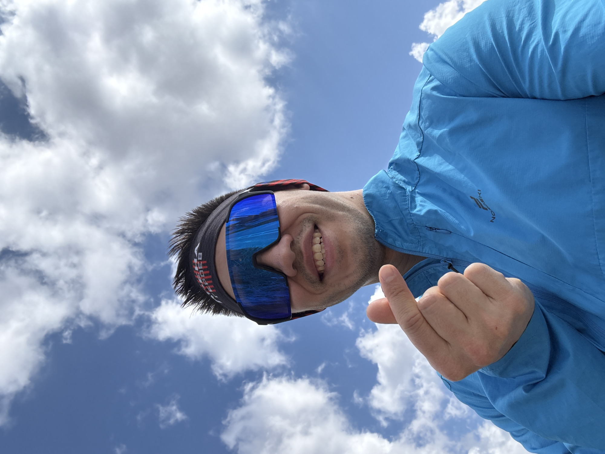 The image is a low-angle, rotated selfie featuring a man looking up at the camera against a bright blue sky. He is smiling widely, revealing his teeth, and his expression conveys cheerfulness and enjoyment. The man wears large, reflective blue mirrored sports sunglasses that cover most of his eyes, and a dark headband with red and white text, partially visible, resembling "KAREB". He is dressed in a bright blue, lightweight jacket, likely a windbreaker, with a subtle "NORSKI" logo visible on the left chest. His right hand is raised, making a gesture with the thumb and pinky extended, often recognized as a "shaka" sign.
The background is a clear, vibrant blue sky interspersed with scattered, fluffy white cumulus clouds, indicating bright, sunny weather. The low perspective suggests the photo was taken from below the subject, looking upwards, possibly during an outdoor activity. The strong natural lighting points to the image being taken during midday or early afternoon. The scene evokes a sense of positive energy and being outdoors on a pleasant day, with the contextual location being Kazan, Russia.
