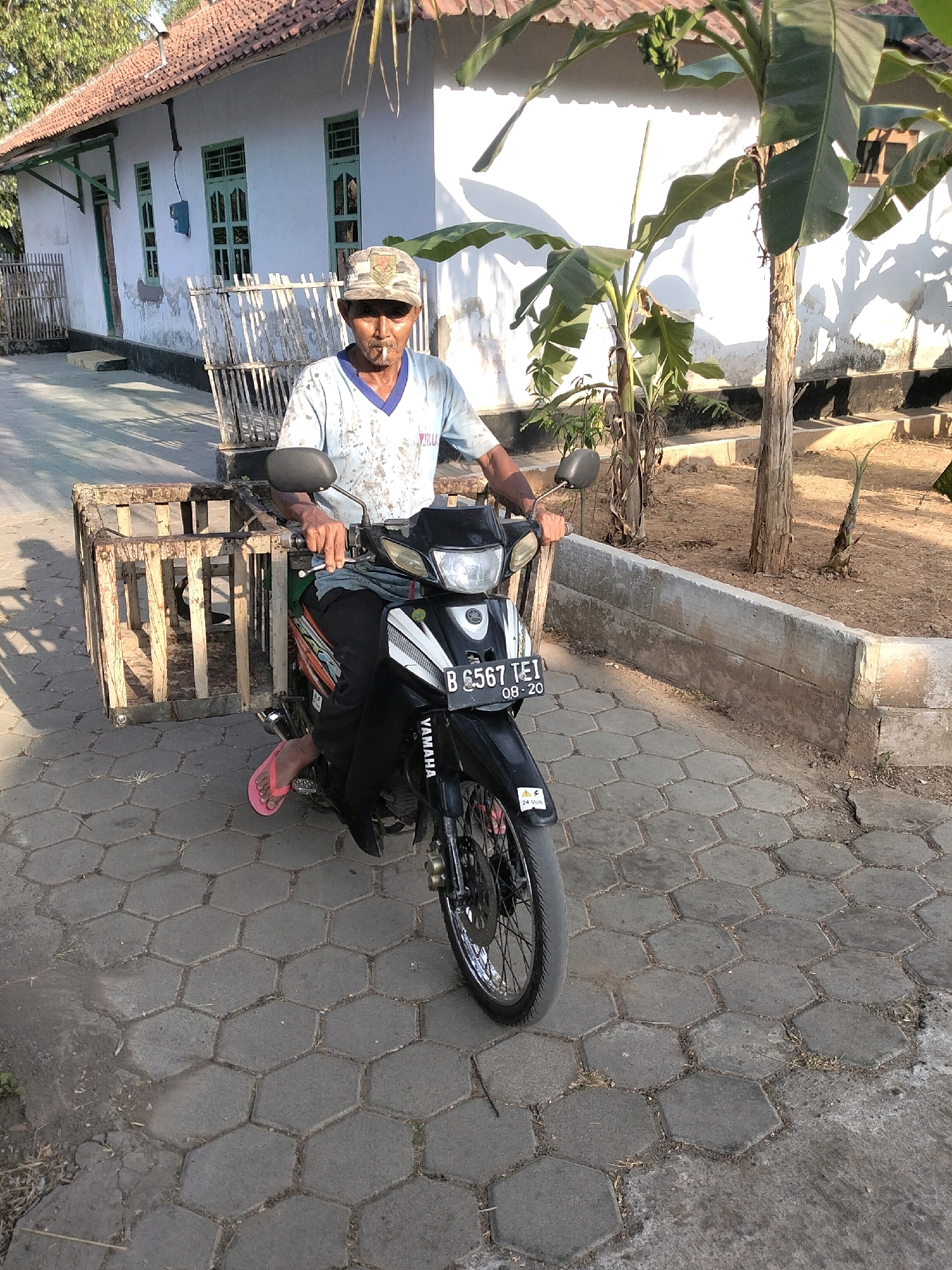 A man with a cigarette in his mouth, wearing a camouflage cap and a light blue shirt with dark accents, sits on a black Yamaha motorcycle. The motorcycle has a license plate that reads "B 6567 TEI" and "08-20". A wooden cart with metal framing is attached to the left side of the motorcycle. The man is wearing pink flip-flops and dark pants. He is on a paved hexagonal-tiled walkway in front of a white building with green-trimmed windows and a red tile roof. Banana trees are visible to the right of the building. The ground is dry and dusty. The scene appears to be in Prapag Kidul, Indonesia, given the context of similar images.