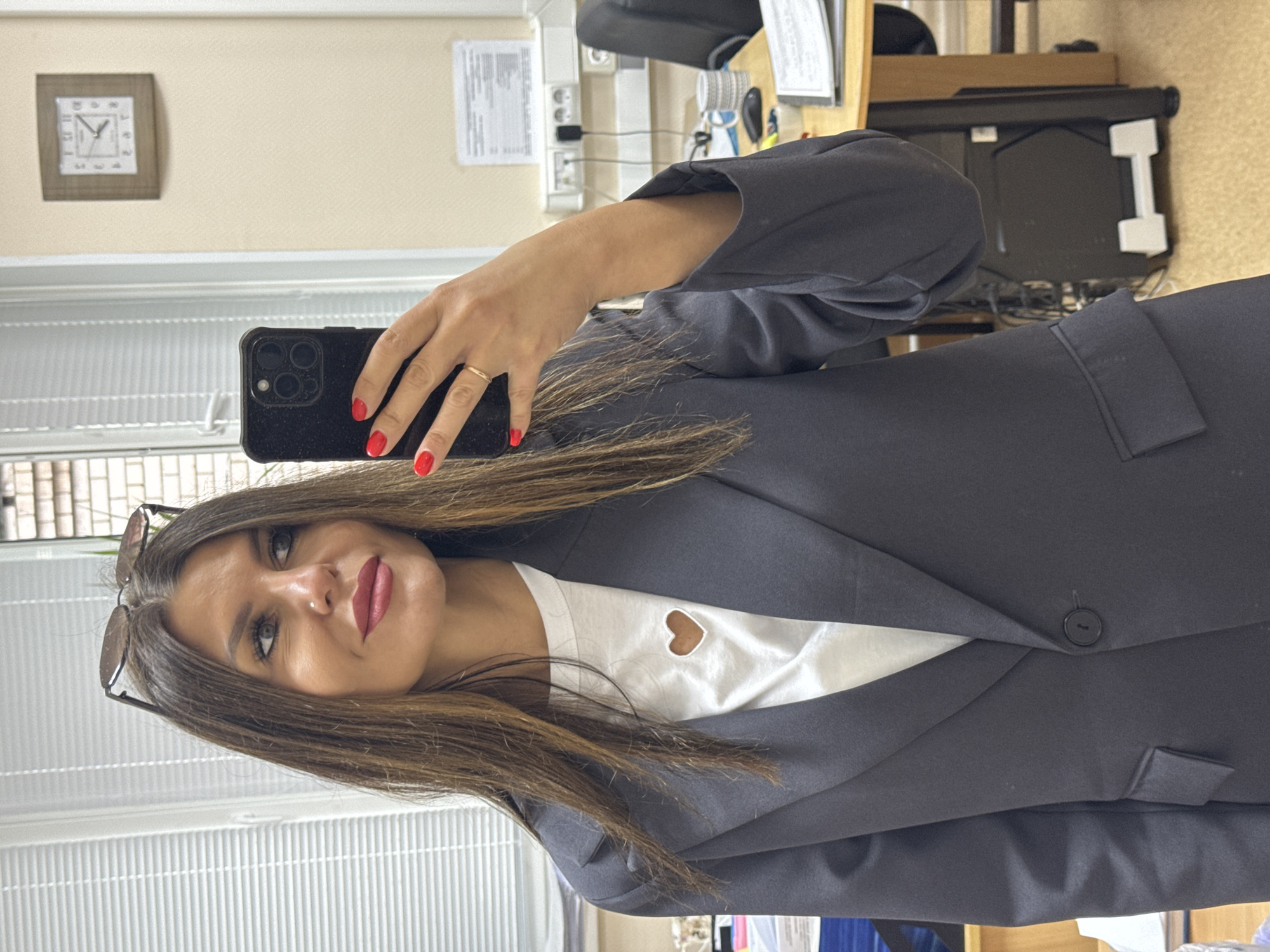 A young woman is captured in a mirror selfie taken within what appears to be an office setting. The image is oriented vertically, suggesting she is holding her phone to capture her reflection.
The woman has long, dark brown hair that falls over her shoulders. She is wearing a dark, possibly navy or charcoal, blazer over a white t-shirt. A notable detail on her t-shirt is a small, light brown heart-shaped cutout on the chest. Sunglasses are perched on top of her head. She has a pleasant, soft smile, with red lipstick and dark eye makeup. A subtle nose piercing is visible. Her left hand, adorned with bright red nail polish and a simple gold ring on her ring finger, holds up a dark-colored smartphone with multiple cameras, possibly an iPhone, towards the mirror.
The background reflects a typical office environment. To the left, a window with vertical blinds is visible. A light-colored wall features a square wooden-framed clock displaying approximately 10:15 or 10:17. Further into the office space, parts of a desk, a black office chair, power outlets, and various electronic equipment and wires are visible. Documents are also seen on the wall and desk. The overall lighting suggests an indoor setting, likely during the morning given the time on the clock. No specific text is legible other than the numbers and possibly "QUARTZ" on the clock face.