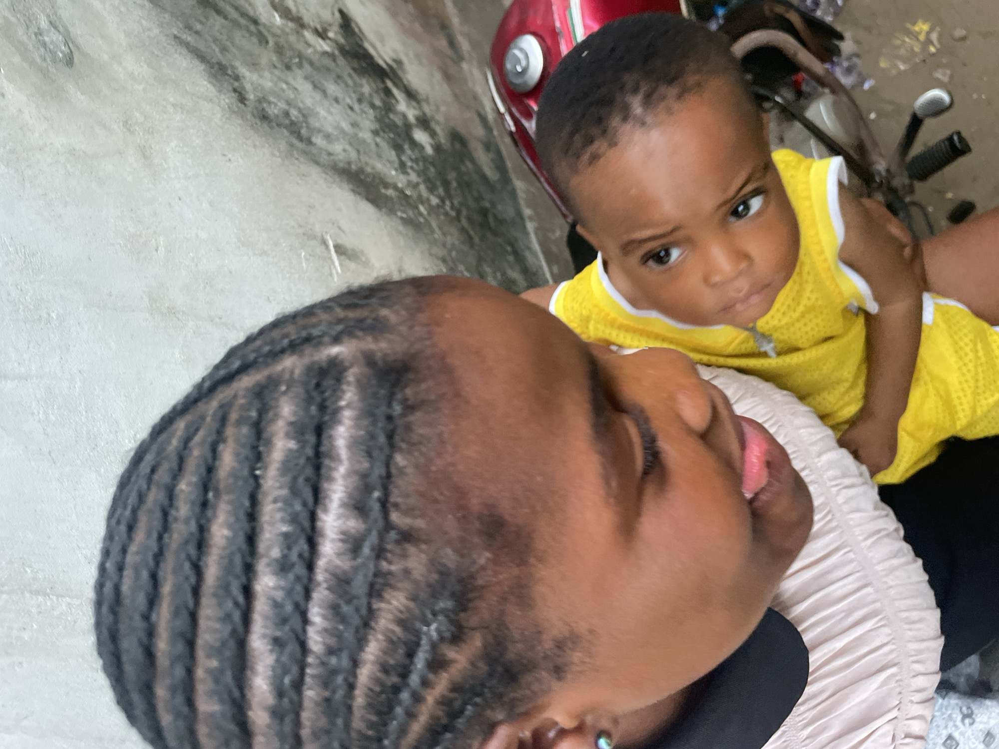 The image captures a close-up, high-angle perspective of a woman with braided hair lying down, with a baby in a yellow outfit positioned above her. The woman's face is turned upwards, with her tongue slightly protruding. The baby looks at the camera with a serious expression. The background includes a red motorcycle and a textured, weathered wall. The location is Badagry, Nigeria.