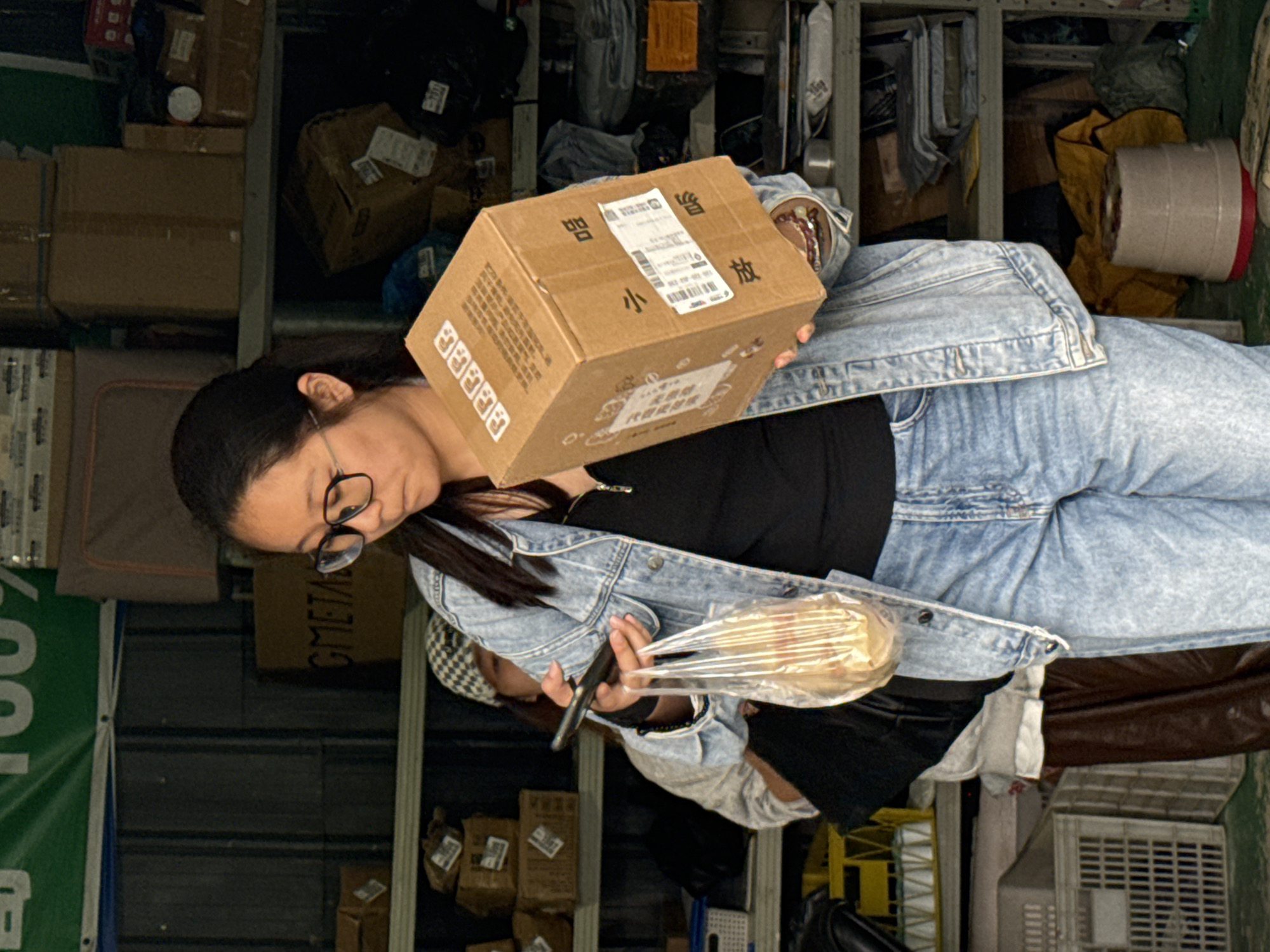 A woman with dark hair and glasses is standing in what appears to be a storage or delivery area. She's holding a cardboard box with Chinese characters on it in her left hand and a phone in her right. She is wearing a denim jacket and a black shirt. The background includes stacked boxes, shelves, and a green banner. Given the context clues of Chinese characters and the user's instructions, the location is likely in Jiangjin, China.