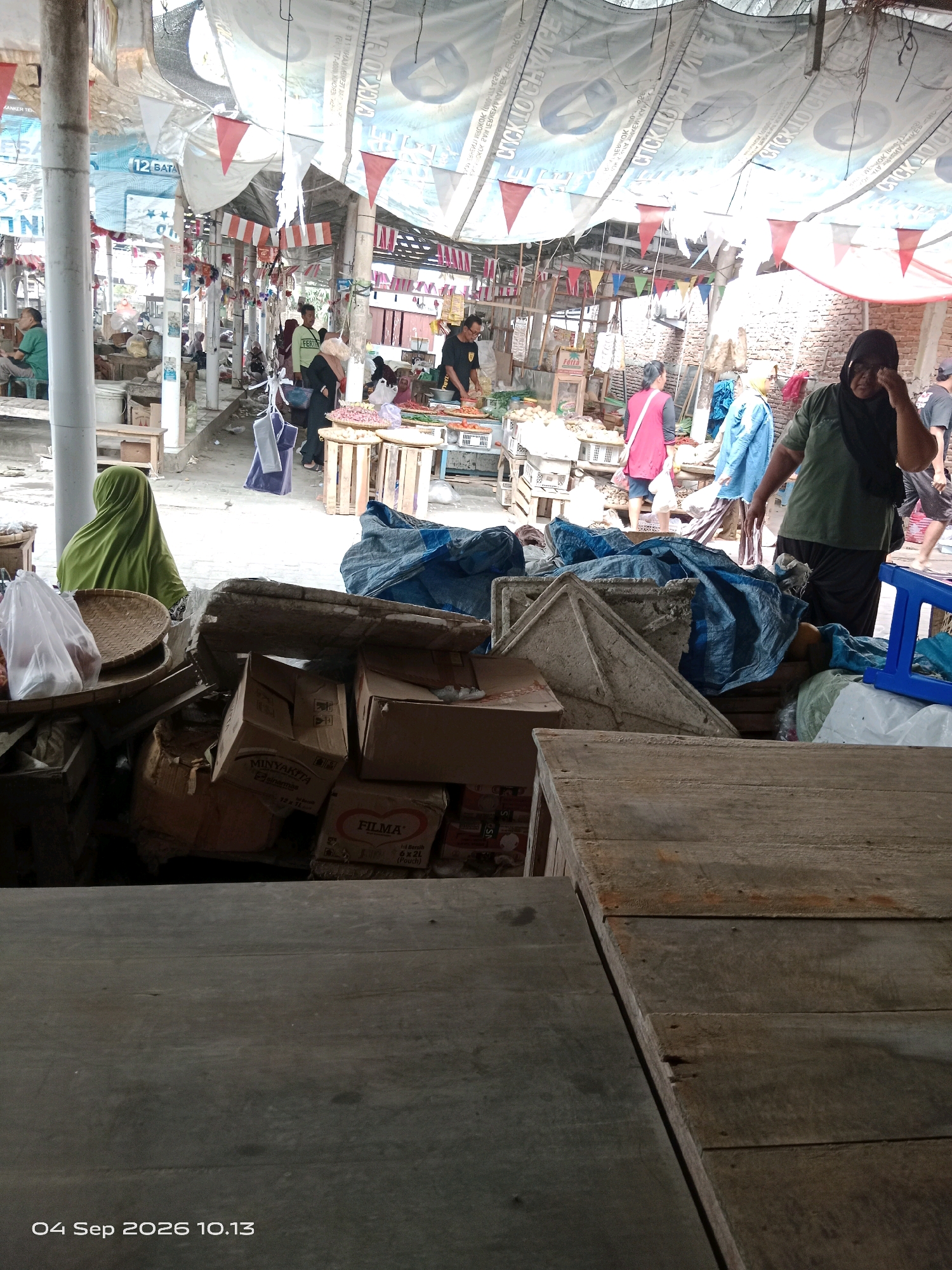 The image depicts a bustling marketplace in Kalipurwo, Indonesia, possibly on a warm day given the attire of the individuals. In the foreground, a wooden surface dominates, with planks showing signs of wear and tear. Behind this, a chaotic arrangement of stacked cardboard boxes and various goods is visible, some covered with blue tarpaulin. One box prominently displays the brand "FILMA." To the left, a woman wearing a green hijab is seen from behind, looking towards the market.
Further into the market, several people are engaged in activities typical of a marketplace. A vendor in a green shirt is interacting with customers, while others are browsing or moving through the stalls. There are displays of produce, including onions and possibly other vegetables and fruits, presented in wooden crates and woven baskets. Colorful triangular flags and banners hang overhead, strung across makeshift stalls, suggesting a festive or celebratory atmosphere. The market appears to be covered by a large tarp, offering shade from the sun. Brick walls are visible in the background, hinting at the surrounding architecture. The overall impression is one of a vibrant, local trading hub filled with activity and commerce. The date stamp at the bottom left corner indicates "04 Sep 2026 10.13".