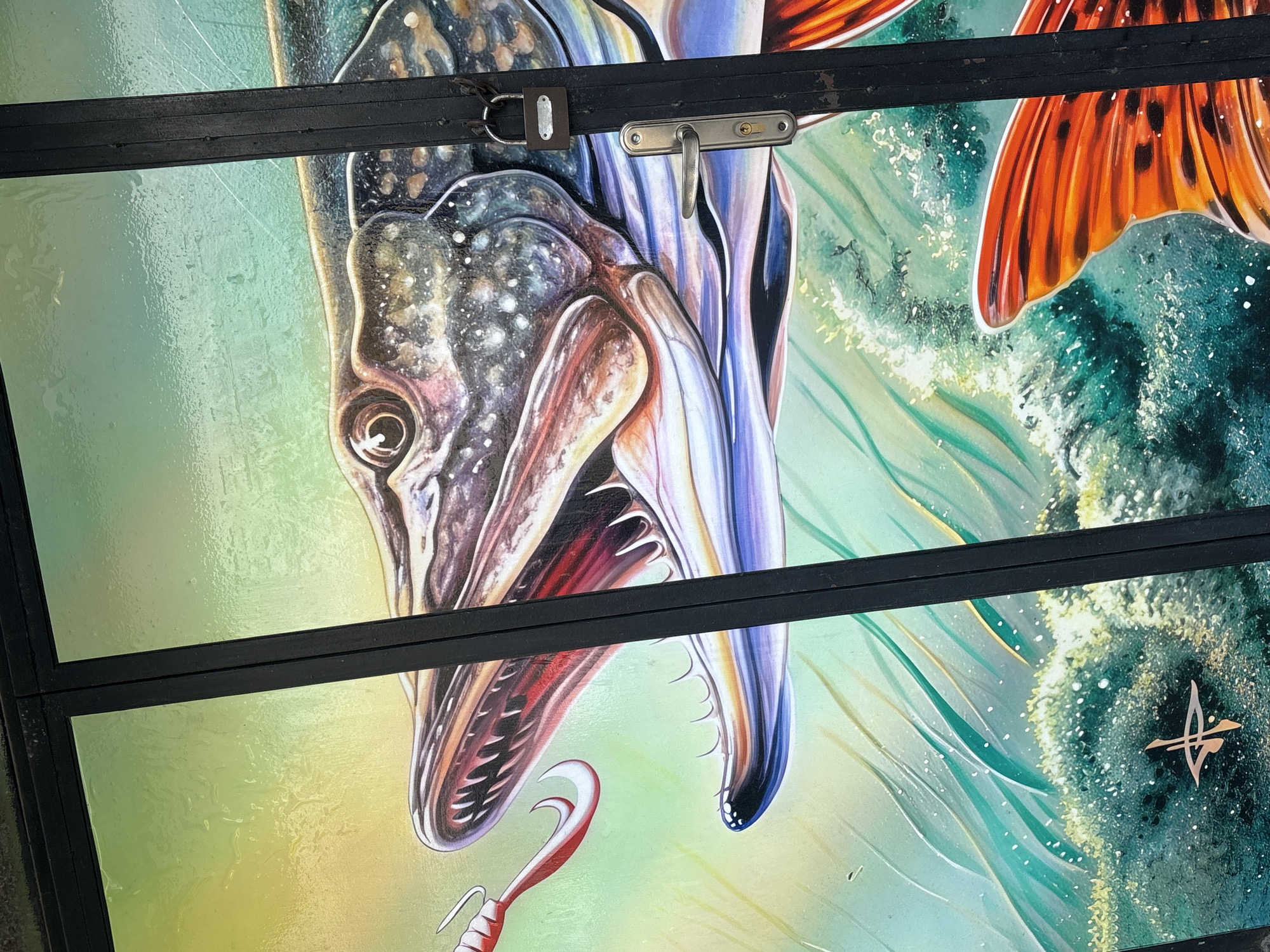 The image displays a vibrant and detailed artwork of a large predatory fish, likely a Northern Pike or Muskie, rendered on a panel structure, possibly a garage door. The artwork is divided by two prominent black horizontal bars, which appear to be structural components of the door.
The central subject is the fish, depicted in a dynamic, near-vertical orientation, with its head and widely open mouth facing towards the viewer from the upper left. Its sharp, conical teeth are clearly visible, along with its detailed eye and speckled scales in shades of grey, blue, and brown. A red and white fishing lure with a hook is positioned near the fish's open mouth, suggesting an active hunting or striking scene. The fish's body and fins extend downwards, with a striking orange tail fin visible on the right side of the image.
The background transitions from lighter green and yellow tones on the left to deeper blues and greens on the right, representing water with splashes and foamy textures, which enhances the sense of movement and action.
Notable details include a silver padlock attached to the upper black bar on the left, and a polished metallic door handle with a keyhole on the same bar, indicating the functional nature of the panel. In the bottom right portion of the artwork, a small, metallic-colored, stylized emblem resembling a bird in flight or a symbolic motif is visible, likely a signature or brand. The surface of the artwork appears glossy or laminated. The scene captures a powerful moment of nature within an urban setting in Plovdiv, Bulgaria.