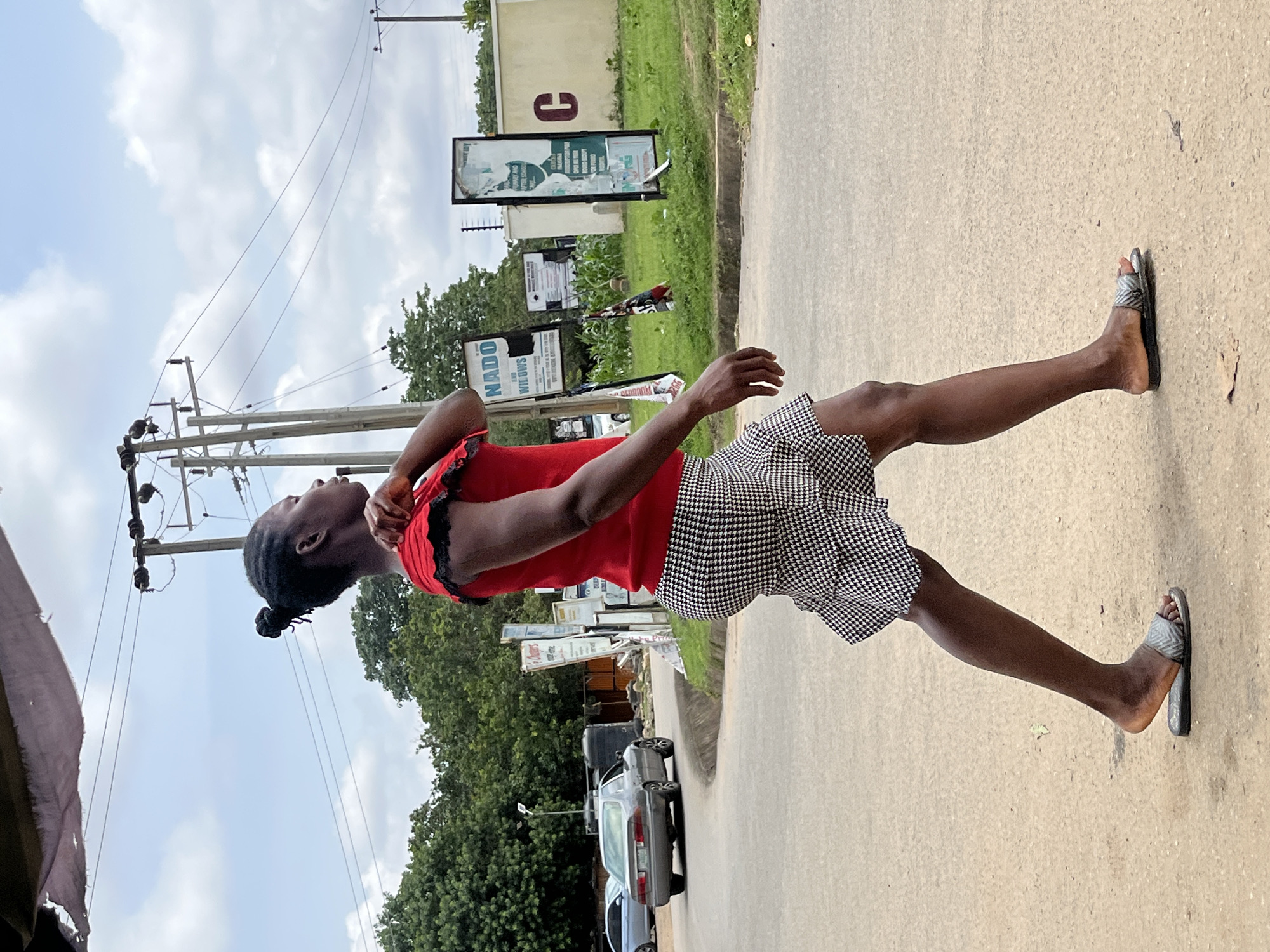A young Black person is standing outdoors on a paved road. They are wearing a red top with black trim and a black and white checkered skirt. They have their hair pulled back in a bun. The person is looking up and to the left, with their right arm raised and bent at the elbow. Their left arm is down by their side. They are wearing sandals on their feet.
The scene appears to be a roadside in Abuja, Nigeria. There are utility poles and wires in the background, as well as some trees and buildings. Several signs are visible, including one that says "NADO". There are also a couple of cars parked on the side of the road. The sky is cloudy, suggesting it might be a partly cloudy day. The lighting indicates it is daytime, but the specific time is not clear. The person's posture and gaze suggest they are observing something above or in the distance.
