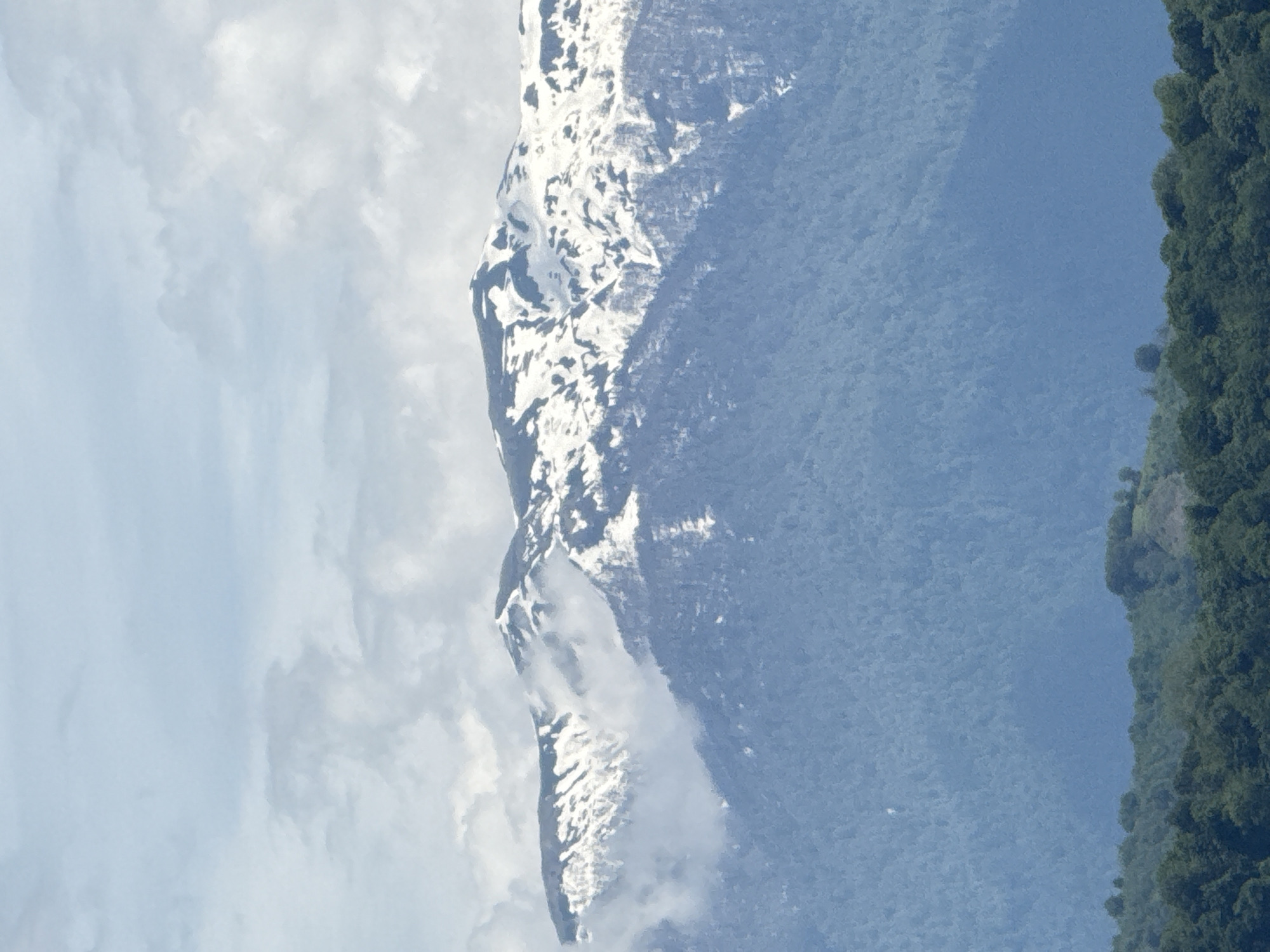 The image shows a mountain in the Krasnaya Polyana region of Sochi, Russia. The mountain has snow on its peak and a forest at its base. The sky is cloudy.