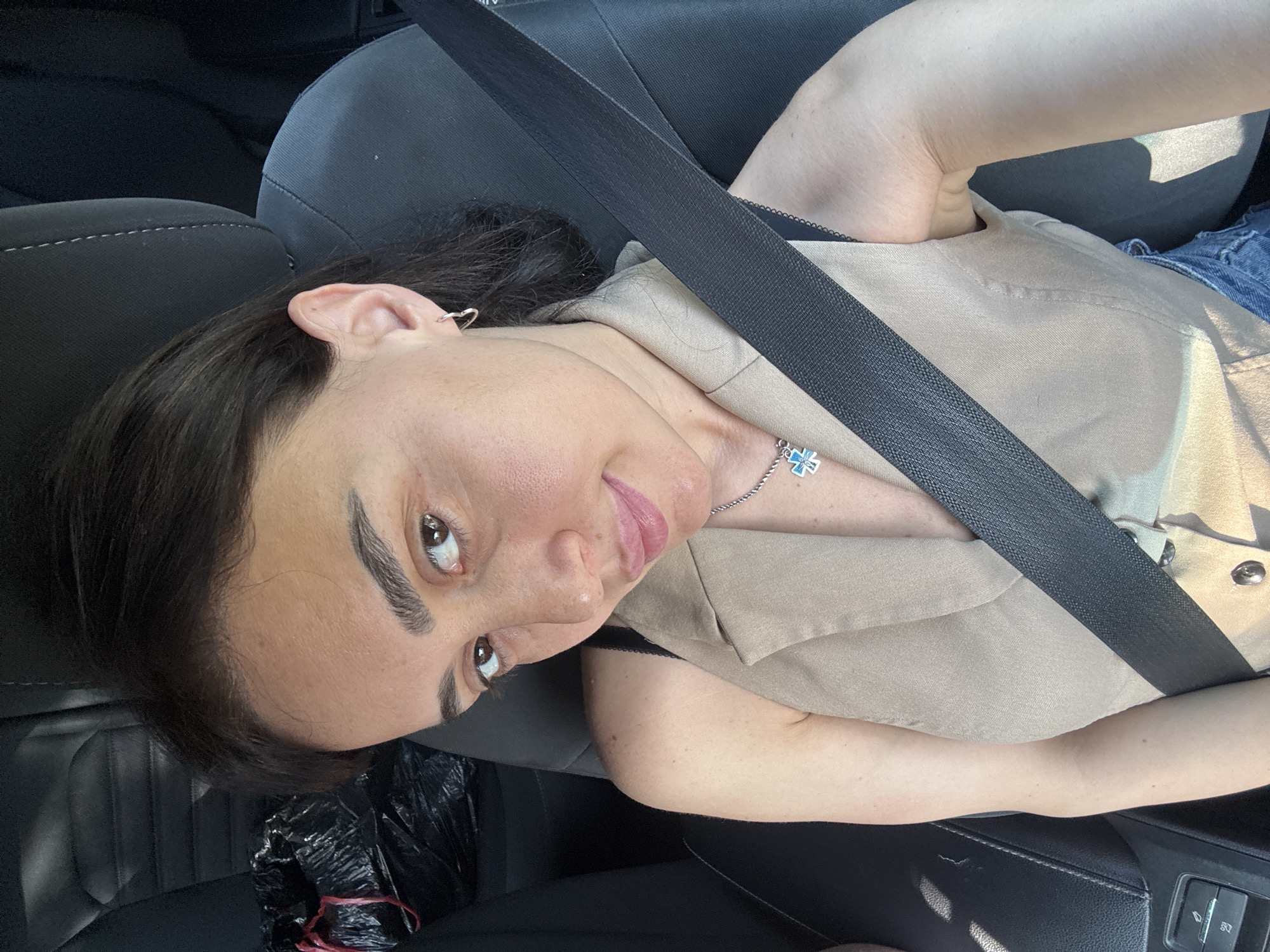 A woman with dark hair and brown eyes is sitting in the driver's seat of a car, looking at the camera. She is wearing a beige sleeveless vest over a black top, and a seatbelt is across her chest. She has a silver necklace with a blue pendant. The car's interior is visible, with a black car seat and a black bag in the background. The image appears to be taken during the day, with natural light coming from the right side of the frame. The specific location in Tbilisi, Georgia is not determinable from the image.