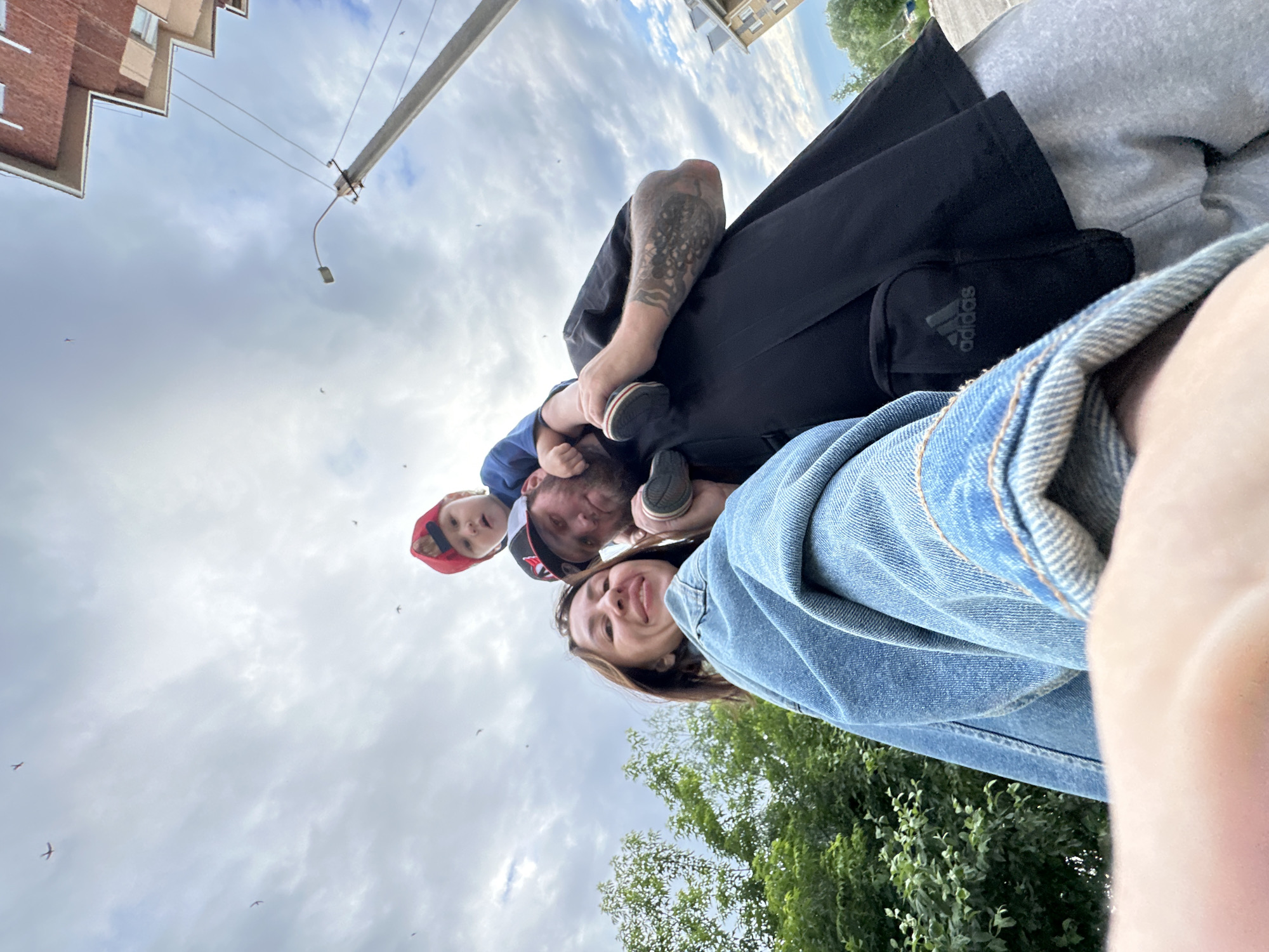 The image is a selfie taken outdoors, with a low-angle perspective looking upwards towards a cloudy sky. In the foreground, a woman wearing a denim jacket is smiling. Behind her, a man is carrying a young child on his shoulders. The child is wearing a red and blue baseball cap. The man has tattoos on his arm and is wearing a black t-shirt with an Adidas logo visible on a small pouch attached to his waist. The background shows greenery from trees, a utility pole with wires, and the corner of a multi-story brick building. Several small dark shapes, possibly birds or insects, are visible in the sky. The overall impression is a casual outdoor family moment.
The provided metadata states the image was taken in Votkinsk, Russia. Considering the visual elements, the scene depicts a typical urban or suburban environment, likely a park, street, or residential area within Votkinsk. The presence of trees, a building, and utility infrastructure are consistent with such a location. The cloudy sky suggests a temperate weather condition, common in Russia.