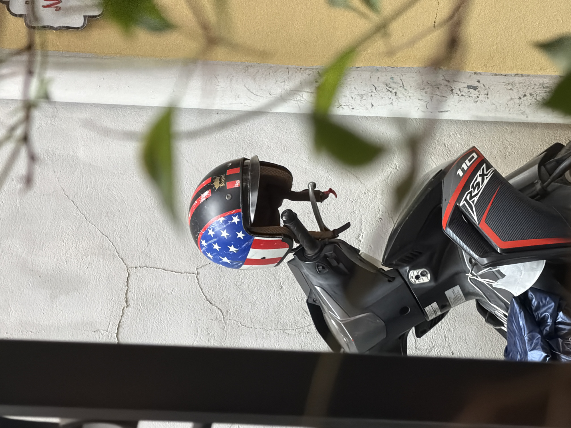 An American flag-themed helmet rests on the handlebars of a parked scooter in Thuận Hóa Ward, Vietnam. The scooter is black with red and silver accents, featuring "RSX 110" branding. A white face mask hangs from the handlebars. The scooter is positioned against a textured, off-white wall with visible cracks. To the right of the wall is a yellow structure. Green leaves are blurred in the foreground and background, obscuring parts of the scene. A dark vertical element, possibly a fence or railing, is present on the left side of the image.