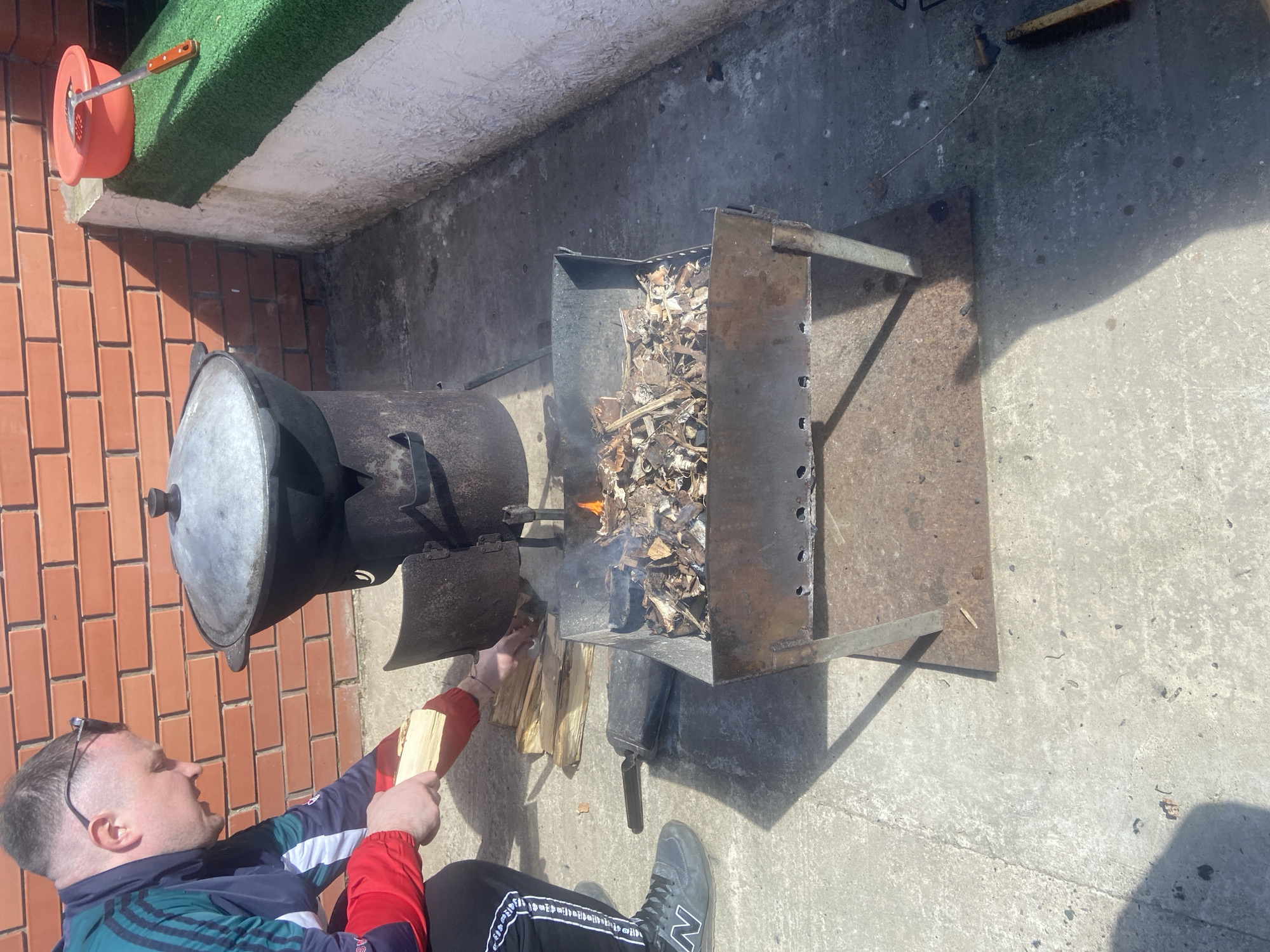A man is adding wood to a fire in a metal container to heat a large, dark-colored cauldron, situated outdoors on a concrete surface. The container is filled with burning wood chips and small flames are visible. The cauldron rests on a stove-like structure directly connected to the metal container.
The man is wearing a dark blue, red, and green jacket with black athletic pants and grey sneakers. He has sunglasses perched on his head.
In the background, there is a brick wall topped with a strip of artificial turf. A red container with a utensil inside is attached to the wall. The setting appears to be outdoors in Ольховка, Russia, based on the presence of Russian text on the man's pants.