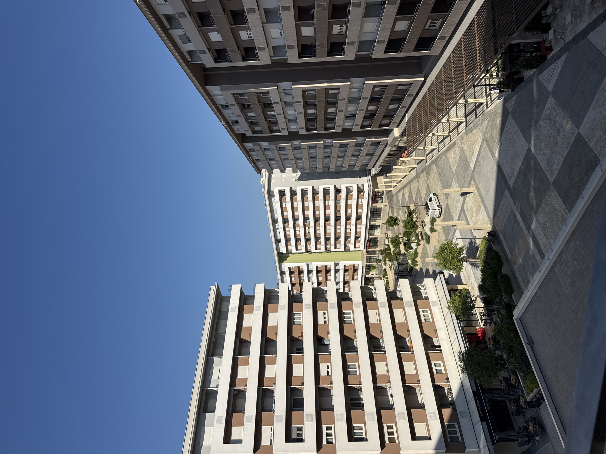 The image shows a modern urban courtyard surrounded by several multi-story buildings. The buildings feature a combination of white, brown, and gray facades with numerous balconies and windows. The courtyard itself is paved with a checkered pattern of light and dark gray squares. Trees and green spaces are interspersed throughout the courtyard. There are also outdoor seating areas for cafes or restaurants. A white van is parked in the courtyard, and a bicycle is leaning against a pole. The sky is clear and blue. The scene is well-lit, suggesting it is daytime.