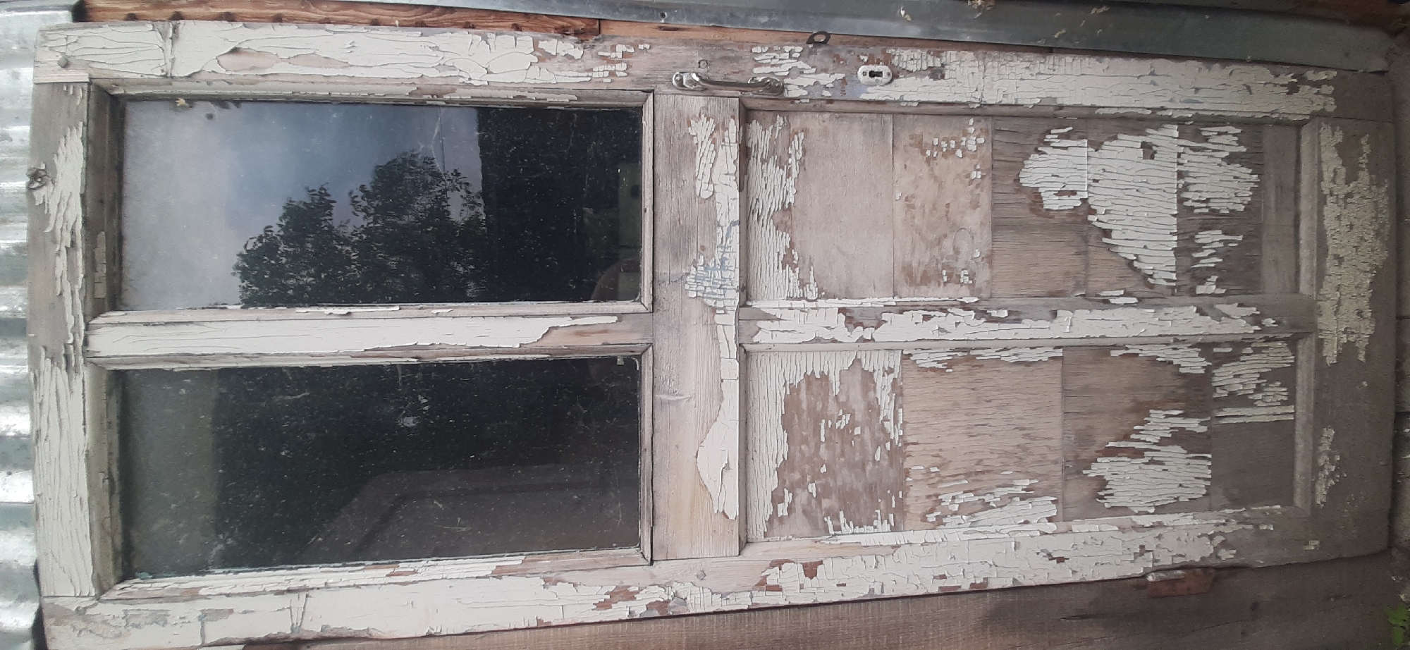 An old, weathered, wooden door with peeling white paint is positioned outdoors, leaning against a corrugated metal surface. The door has two glass panes at the top, reflecting trees and a cloudy sky. The lower half of the door is paneled wood, also with significant paint loss, revealing the underlying wood grain. A metal door handle and a keyhole are visible on the right side. The background shows a wooden wall to the left and the corrugated metal structure to the right. The scene is likely in Yessentukskaya, Russia, given the context.