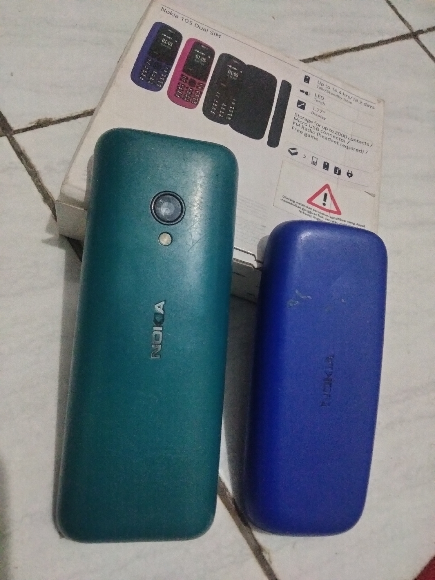 The image displays two Nokia mobile phones, one teal and one blue, placed on a textured surface, possibly a tiled floor in Antirogo, Indonesia. A Nokia 105 Dual SIM phone box is partially visible in the background, showing product specifications and images of the phones. The teal phone is positioned on the left, showcasing its camera and the "NOKIA" branding. The blue phone is on the right, also displaying the "NOKIA" logo. The environment appears to be indoors, with the phones and the box being the primary subjects of the photograph.
