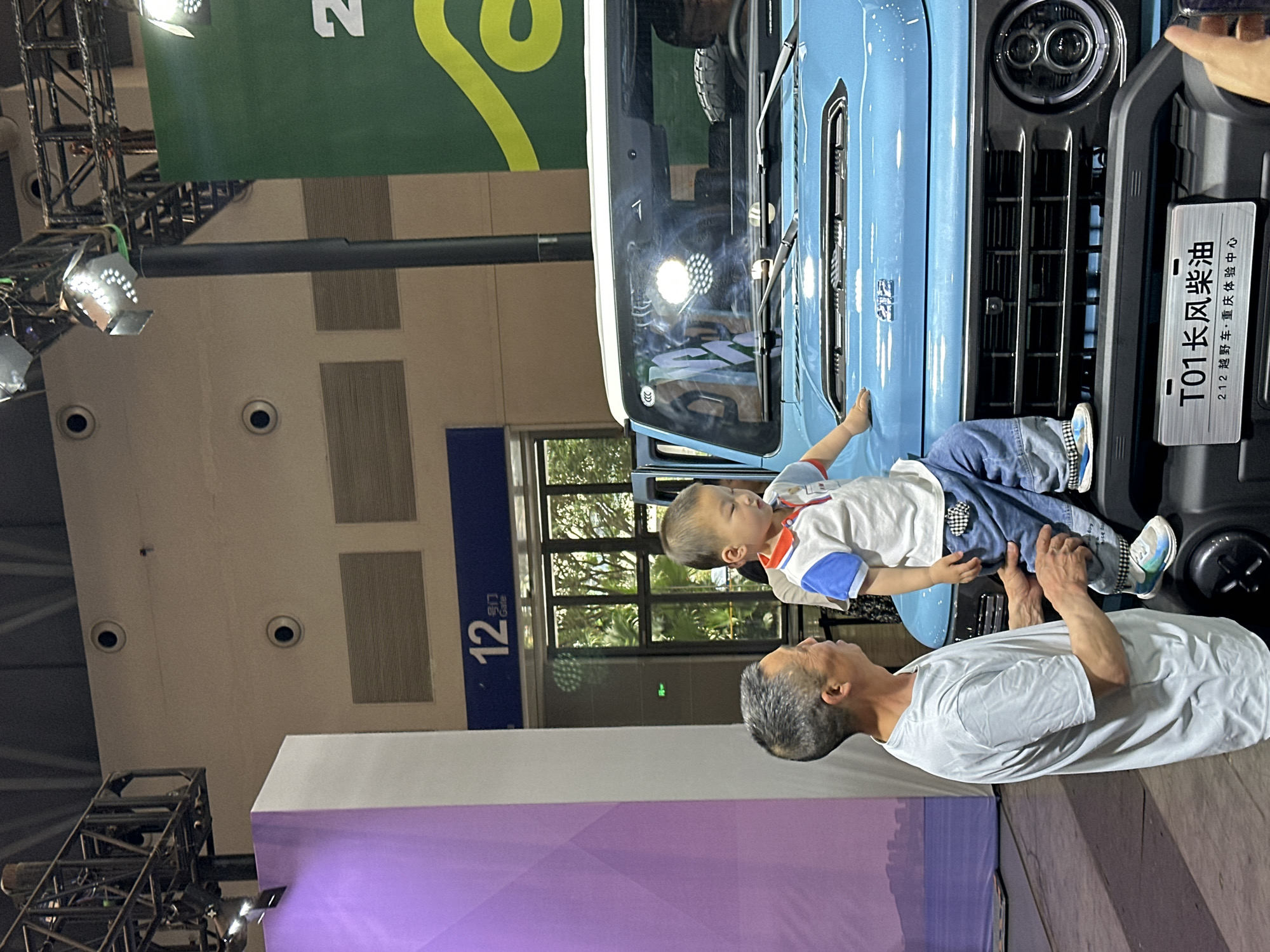 A scene captures a child being held up to a blue vehicle at an automobile expo in Liangjiang, China. The vehicle, marked with "T01长风柴油," appears to be a "212 越野车·重庆体验中心" model. A man, presumed to be an adult, supports the child, who is dressed in a casual outfit. The location is an exhibition center with interior lighting, visible ceiling fixtures, and a partially obscured view through a window in the background.