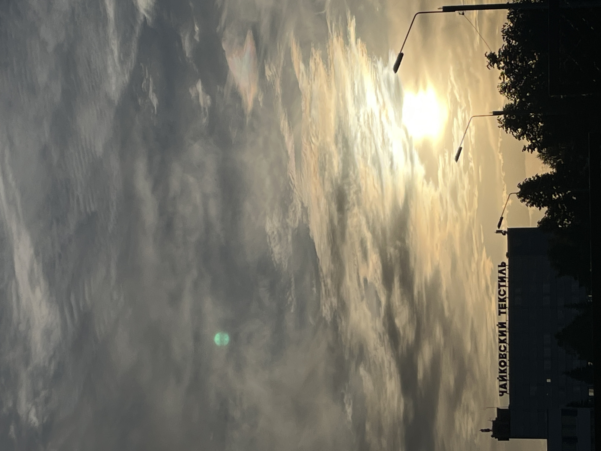 The image depicts the sky over Chaikovsky, Russia, at what appears to be sunrise or sunset, given the position of the sun. The sky is filled with streaky clouds, and the sun is shining through them. A building with the words "Чайковский текстиль" (Chaikovsky Textile) is visible, indicating the location. There are street lamps and silhouettes of trees adjacent to the building. A green lens flare is visible in the lower left portion of the image.