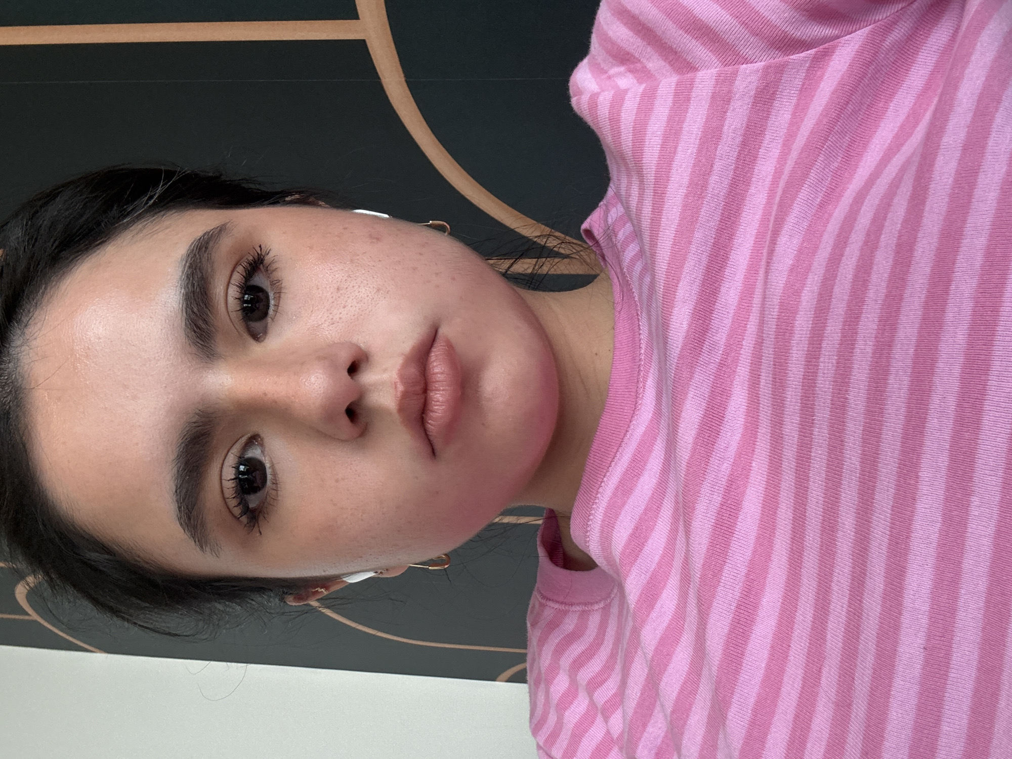 A woman with dark hair and light skin, wearing a pink and white striped shirt and gold hoop earrings, is facing the camera. She is likely indoors, with a wall decoration featuring a dark gray background and a curved gold line visible behind her. The location is impossible to determine from the image alone. The image is rotated to the side.