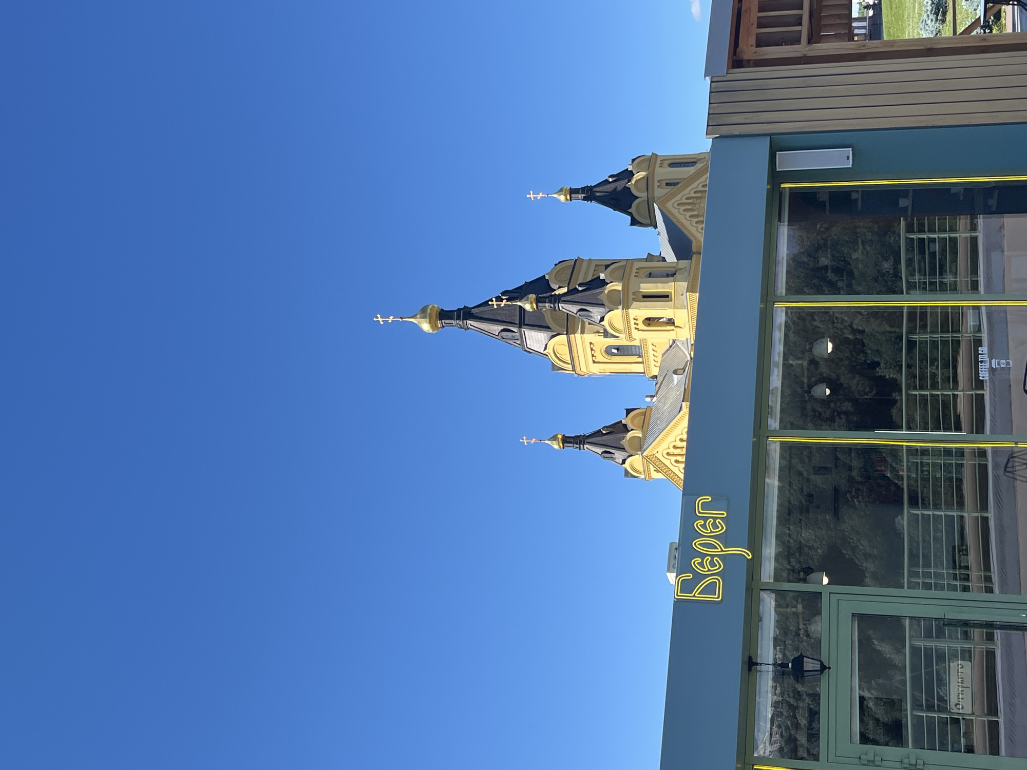 The image depicts a bright, clear day with a vivid blue sky. Dominating the upper portion of the frame are the distinctive onion domes and spires of a yellow and black church, characteristic of Russian Orthodox architecture. The church appears to be a significant landmark, its ornate structure silhouetted against the sky.
In the foreground, a modern building with a teal-colored facade and large glass windows occupies the lower right section. A neon sign in a cursive, yellow font is visible on the teal facade, spelling out "Beper". The windows reflect the surrounding environment, showing glimpses of trees and interior lighting. Green metal framing surrounds the windows and doors, adding a contemporary touch to the building's exterior.
The composition suggests an urban or semi-urban setting, where a historical religious building stands near a more recent commercial structure. The lighting indicates it is daytime, likely mid-morning or afternoon, given the strong sunlight and clear sky. There are no people visible in the image, focusing the viewer's attention on the architectural elements and the sign. The overall impression is one of a sunny day in a location where historical and modern architecture coexist.