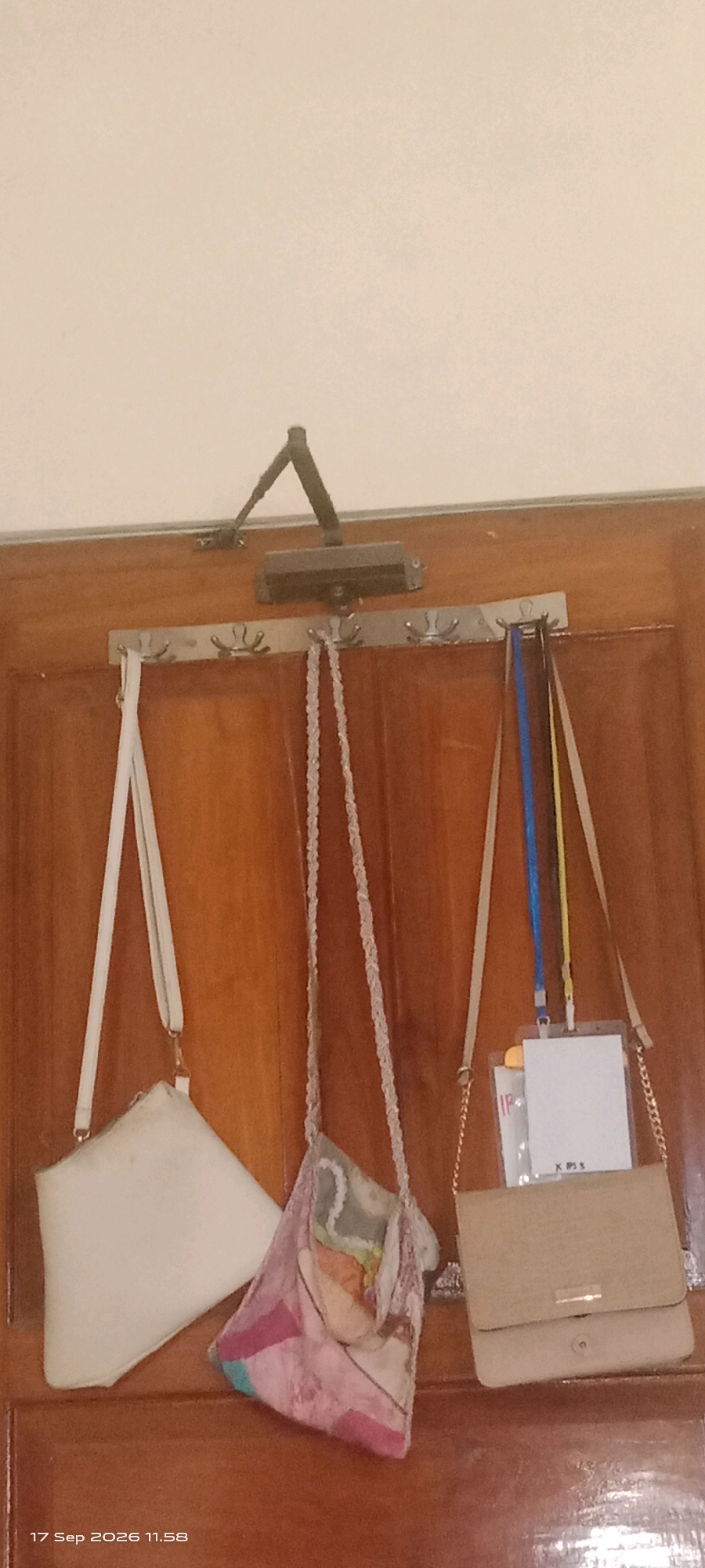 The image shows a wooden door with a metal coat rack mounted horizontally across it. Three bags are hanging from the coat rack. On the left, a light-colored envelope-style handbag is suspended by a thin white strap. In the center, a multi-colored, patchwork fabric bag with a braided strap hangs. To the right, a beige flap-over handbag is attached to a thin chain strap. A lanyard with a clear plastic ID holder is attached to the right side of the beige handbag, with a blue and yellow cord visible. A door closer mechanism is visible mounted above the coat rack on the door frame. The bottom of the image shows a timestamp reading "17 Sep 2026 11.58". The overall setting appears to be indoors, likely a residential or office environment. The wood grain of the door is clearly visible.