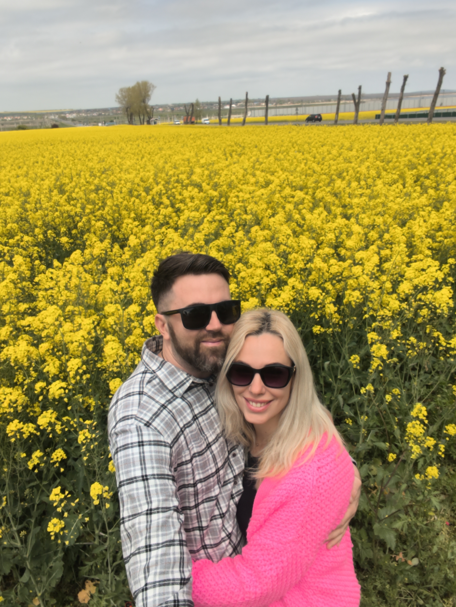 A couple stands embracing in a field of yellow flowers in Ленинское сельское поселение, Ukraine. The man has a beard and wears a plaid shirt and sunglasses. The woman has blonde hair and wears a pink sweater and sunglasses. A road with vehicles and a line of wooden posts is visible in the background. The sky is overcast.