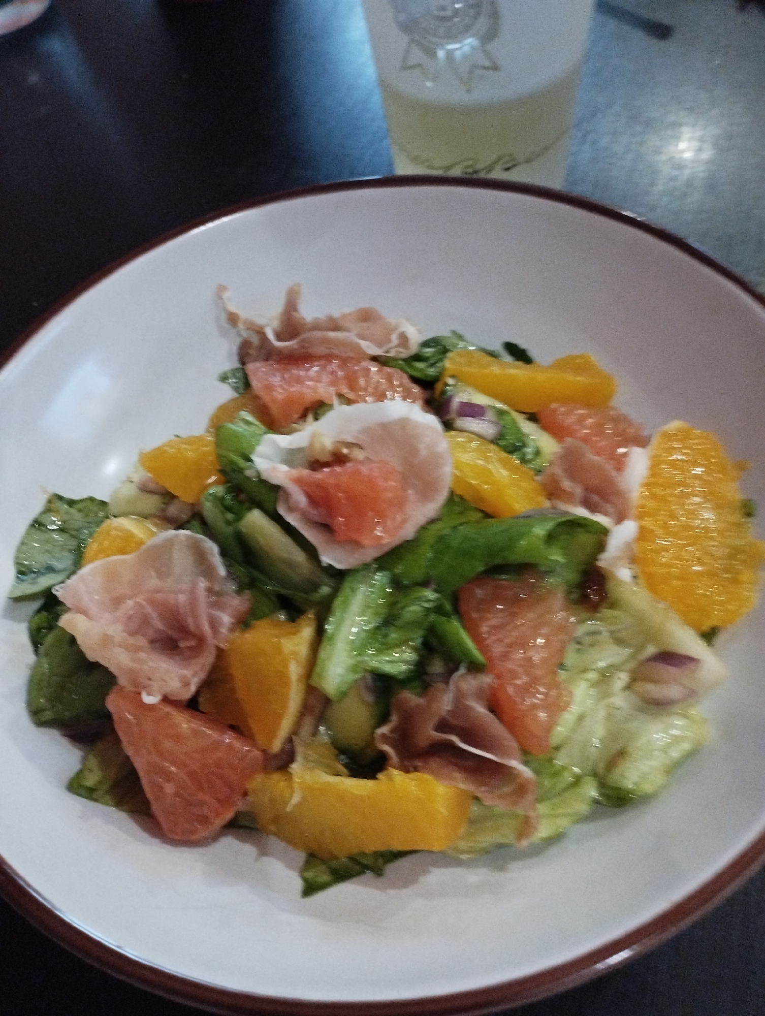 A close-up shot depicts a salad in a white bowl with a brown rim, situated on a dark surface. The salad contains a mixture of lettuce, orange slices, pink grapefruit wedges, and prosciutto slices. A glass with a logo or emblem on it sits slightly out of focus in the background, partially obscuring the upper part of the image. The salad's ingredients appear fresh, and the overall composition suggests it's part of a meal. The setting suggests a restaurant or dining environment. There is no evidence to link the image to the city of Burgas, Bulgaria.