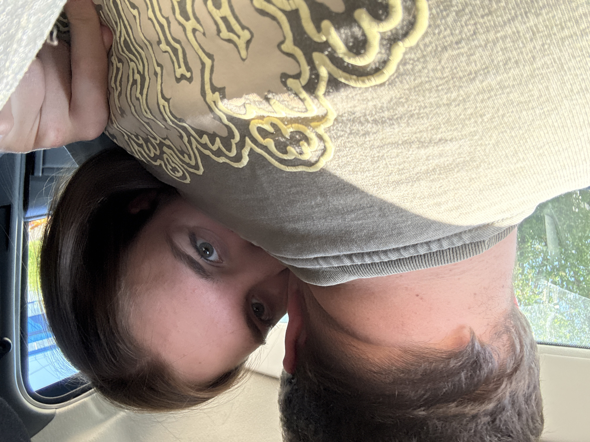 A close-up, upside-down selfie of two people inside a vehicle. One person with fair skin and dark brown hair is angled to the left, looking at the camera. The other person, with dark hair, is positioned to the right. A light-colored fabric or clothing item with a darker-colored decorative design is near the top of the image. A vehicle window is visible in the background, showing green trees and what appears to be a blue sky. No specific landmarks or details indicate the location is Perm, Russia.