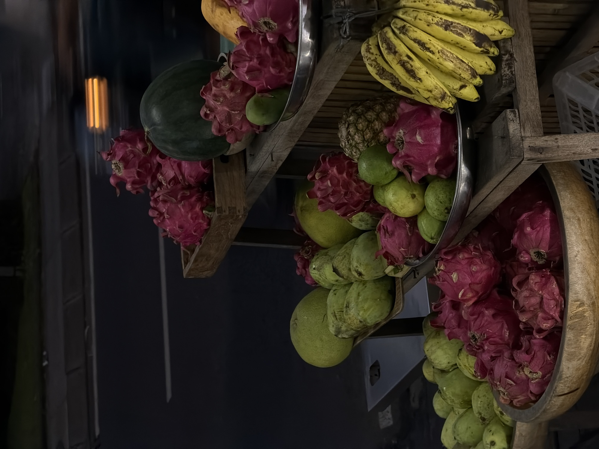 The image shows a fruit stand in North Kuta, Indonesia. The stand is made of wood and has multiple levels. There are various types of fruit on display, including dragon fruit, bananas, mangos, watermelons, and pineapples. The fruit is arranged in baskets and metal trays. The background is dark and out of focus.