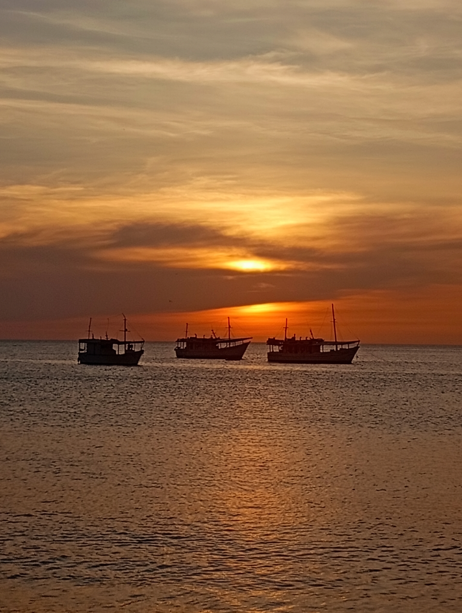 The image captures a sunset scene at Juan Griego, Venezuela. Three boats are positioned on the water, appearing as silhouettes against the golden and orange hues of the sky. The sky is partially covered by clouds, with the sun peeking through. The water reflects the colors of the sunset, creating a shimmering effect.