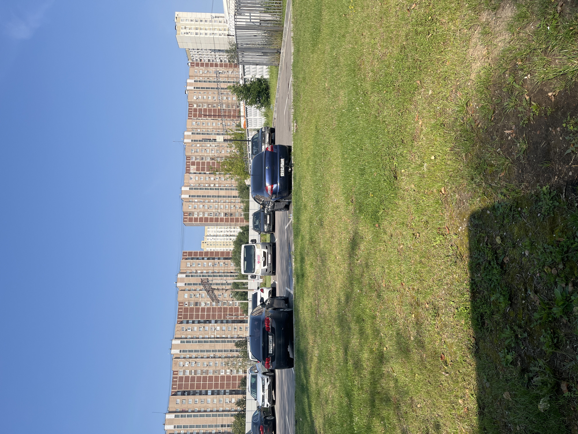 In Moscow, Russia, an outdoor parking lot is visible with several cars and a white van. In the background, a row of tall, tan and red apartment buildings stretches across the horizon under a clear blue sky. The foreground features a patch of well-maintained green grass. The scene is set during the daytime with bright sunlight casting shadows. The environment appears to be a typical urban residential area.