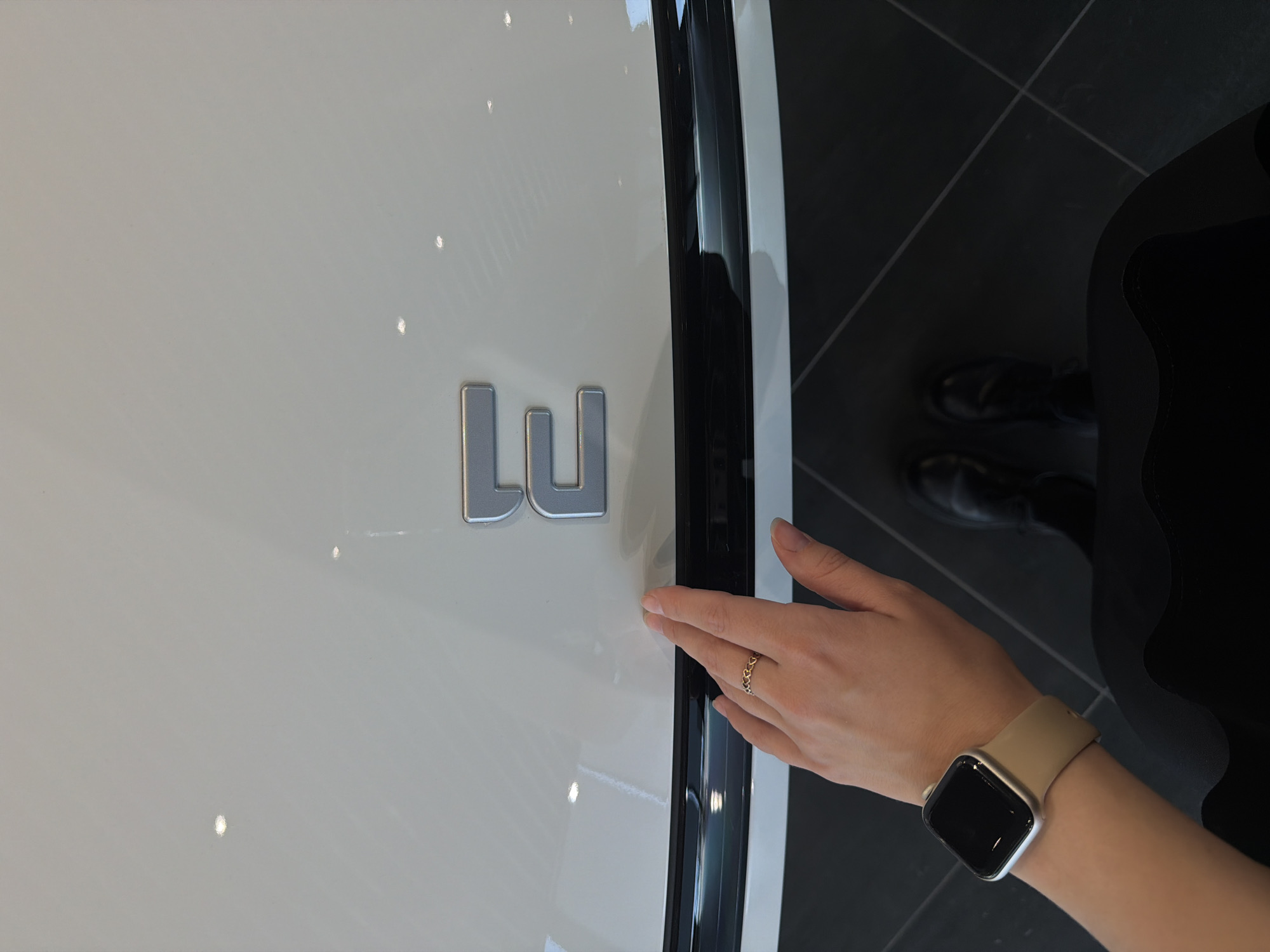 The image shows the front of a white car with a silver "LU" emblem on the hood. A person's hand with a gold ring is touching the edge of the hood. The person is wearing an Apple watch with a beige strap. The background shows a dark tiled floor and the person is wearing black pants and shoes. The location cannot be determined from the image.