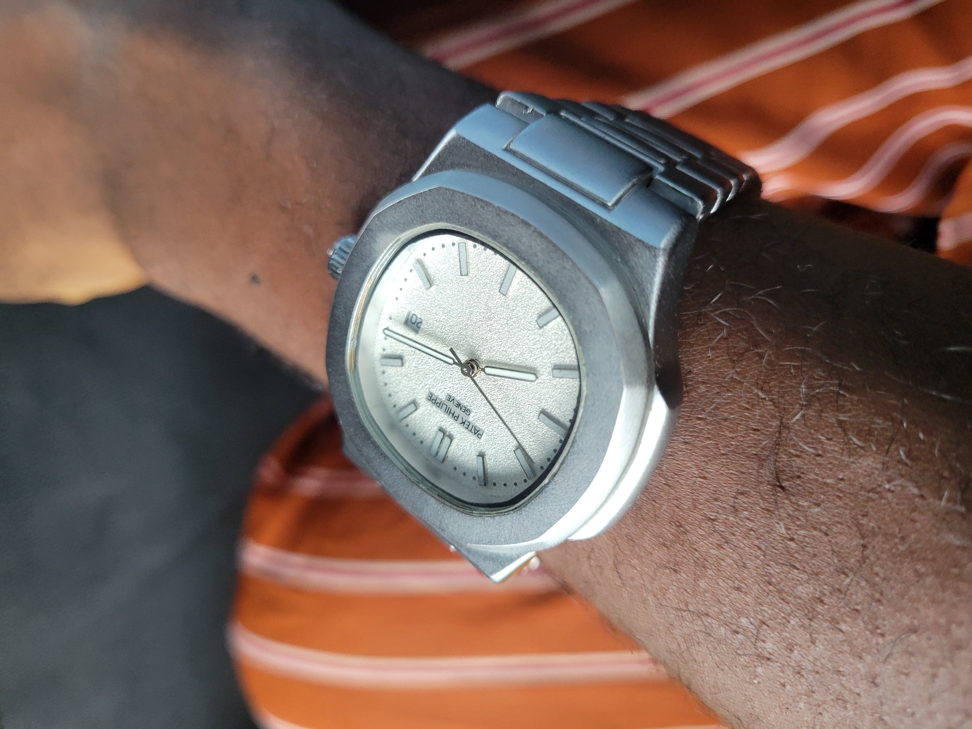 A close-up shot depicts a person's wrist adorned with a silver-toned wristwatch. The watch features a distinctive angular bezel and a light-colored dial with silver hands. The watch band is also silver-toned, appearing to be made of linked metal segments. The person's skin is dark-toned and hair is visible on the arm. The individual is wearing an orange and white striped garment. The location and city are unknown.