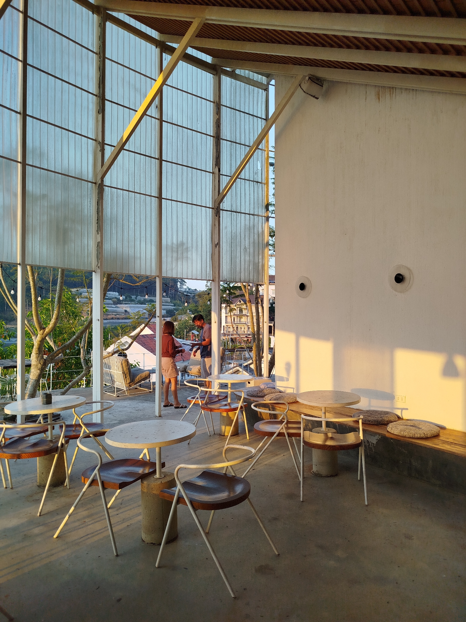 The image depicts the interior of a cafe, presumably located in Da Lat, Vietnam. The space is characterized by a combination of natural light and architectural elements. There are several tables and chairs arranged in the foreground, with a focus on a minimalist aesthetic. A translucent wall composed of vertical panels provides a view to the outside where two people are standing. To the right, a tall, white wall features two circular fixtures. The light source appears to be from the side, casting long shadows across the floor. The overall atmosphere suggests a relaxed, modern setting.