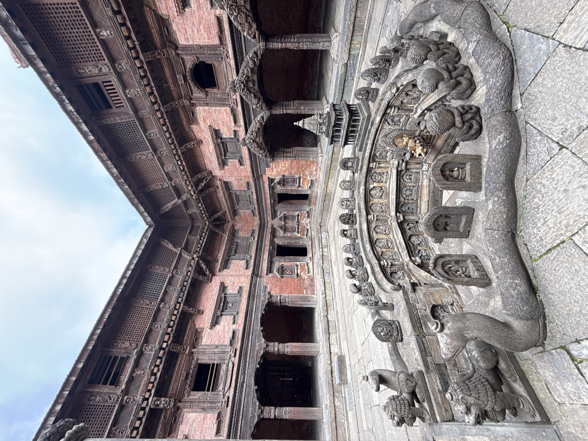 The image shows the architectural details of the Golden Gate at the Patan Durbar Square, located in Lalitpur, Nepal. The gate is elaborately decorated with stone carvings of mythical creatures, including lions and snakes. The building's facade, constructed of brick and wood, features intricate window designs and detailed latticework. Arched entryways are visible, leading into the building. The sky, with patches of blue, is seen in the background.