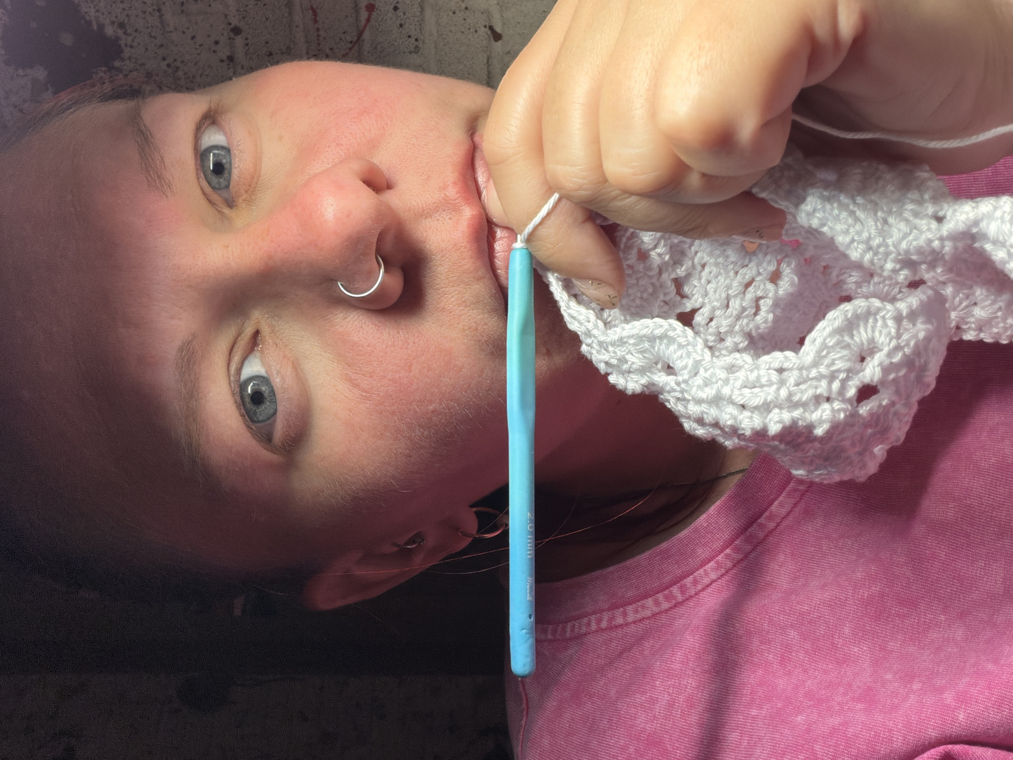 A person is holding a crochet hook with white yarn and a crocheted piece. The person has gray eyes and a nose ring. They are wearing a pink t-shirt. The scene appears to be indoors with a mottled gray background. The city of Chaikovsky, Russia, cannot be verified from the image.